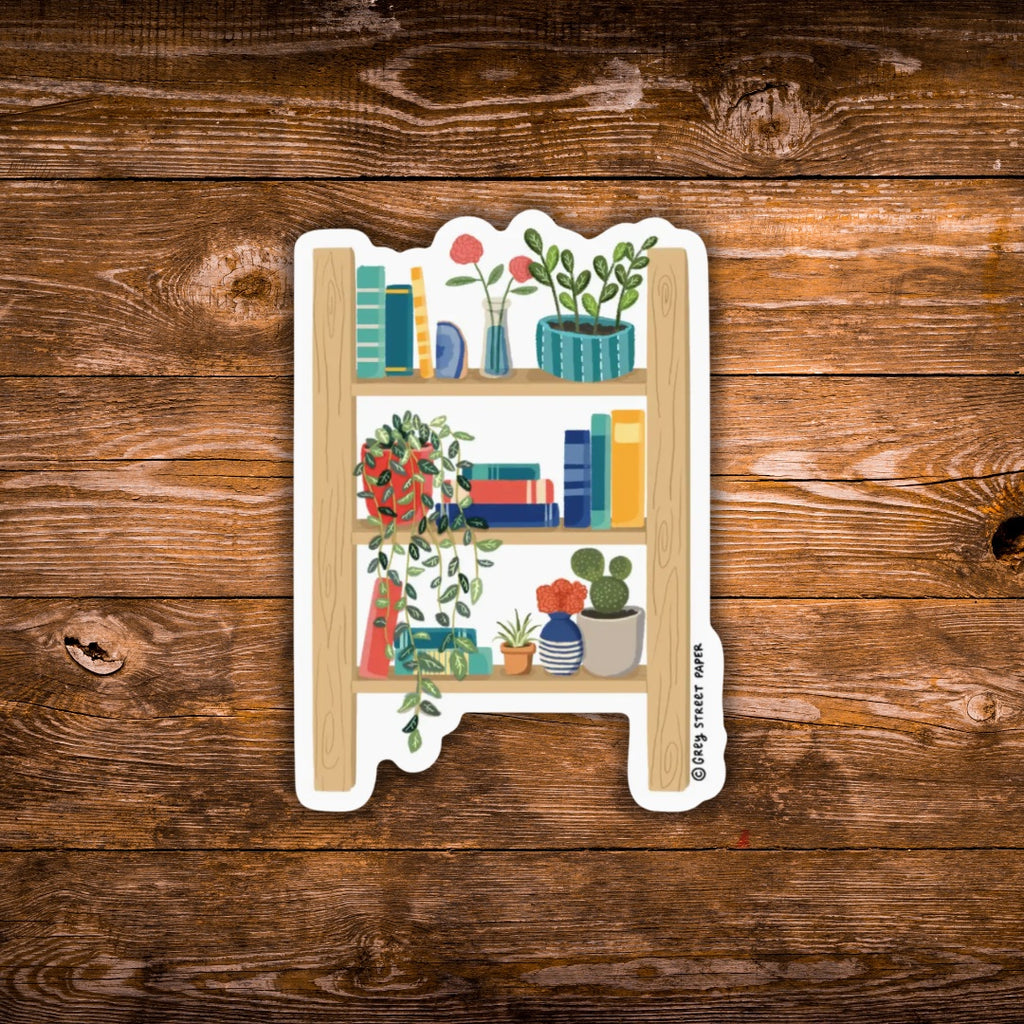 A die-cut sticker featuring a whimsical wooden bookshelf filled with books, plants, and flowers, displayed on a rustic brown wooden background.