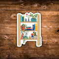 A die-cut sticker featuring a whimsical wooden bookshelf filled with books, plants, and flowers, displayed on a rustic brown wooden background.