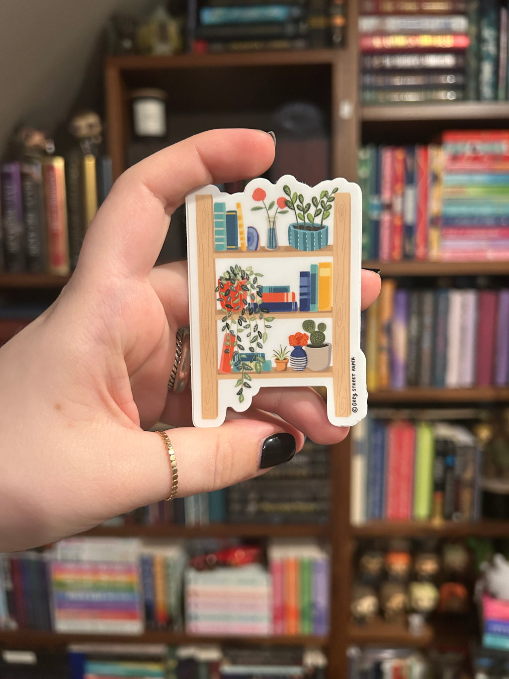 A hand holds a small sticker of a colorful bookshelf illustration. The background is a softly blurred bookshelf.