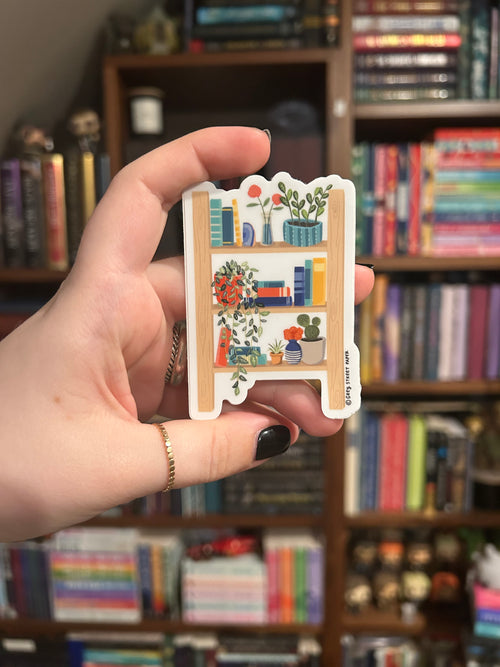 A hand holds a small sticker of a colorful bookshelf illustration. The background is a softly blurred bookshelf.