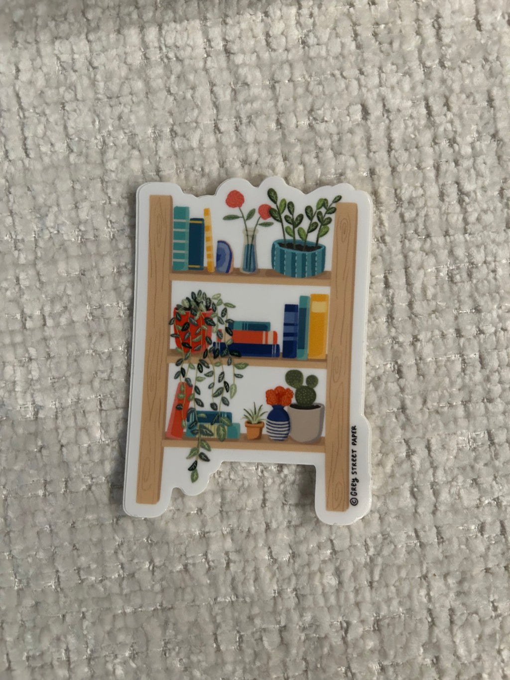 A small decorative sticker with a white border and a colorful illustration of shelves, books, plants, and flowers, placed on a textured light gray fabric background.