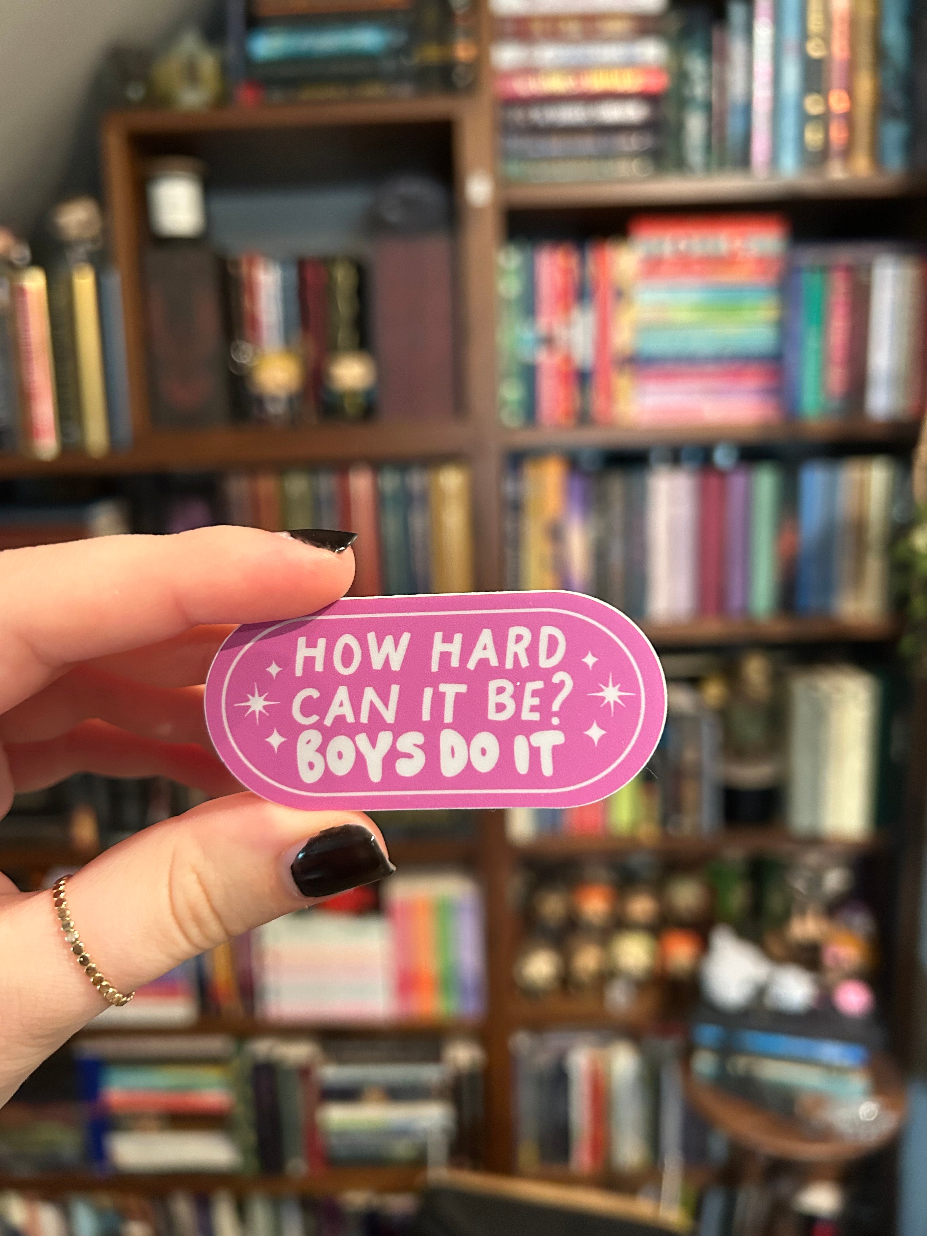 A hand with black nail polish holds a pink oval sticker that reads 'HOW HARD CAN IT BE? BOYS DO IT' against a blurred background of bookshelves.