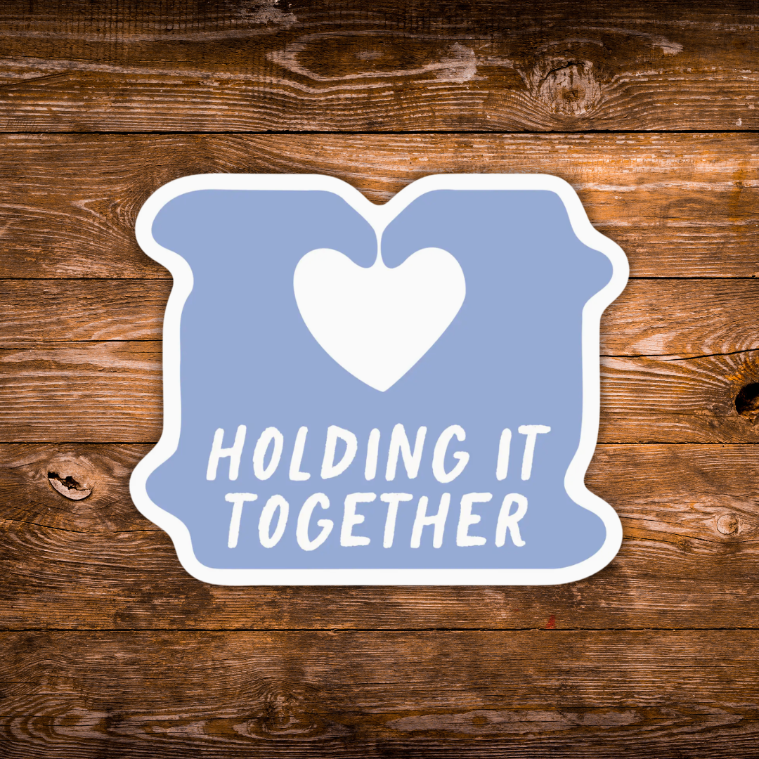 A die-cut sticker with a light blue background, a white heart in the center, and the words 'HOLDING IT TOGETHER' in white. The sticker is displayed flat on a rustic brown wooden plank background.