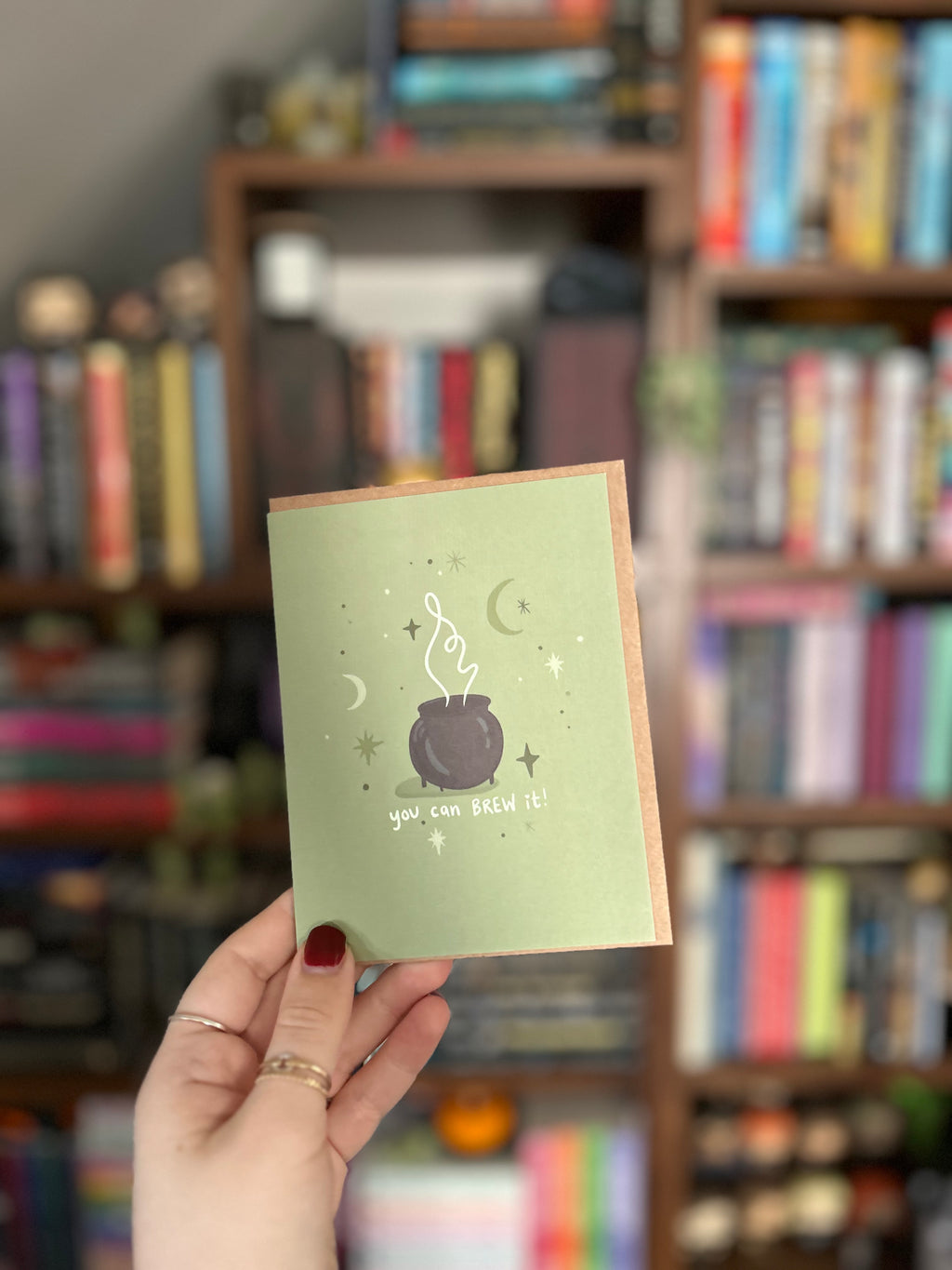 Encouragement greeting card featuring a bubbling cauldron surrounded by stars and crescent moons with the message "You Can Brew It!" on a sage green background.