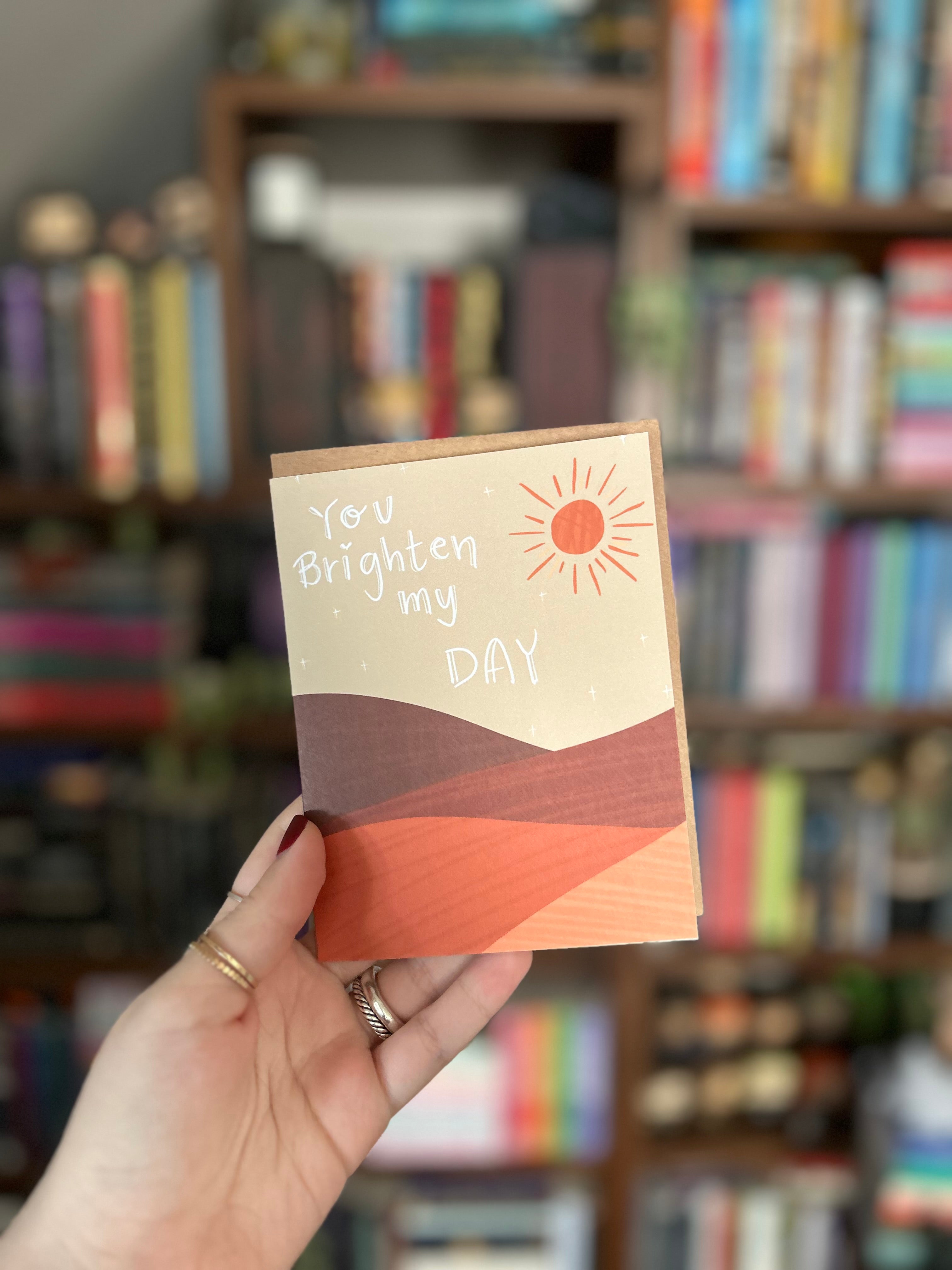 Modern greeting card featuring a warm illustrated landscape with a bright sun and the message "You Brighten My Day" on a soft neutral background.