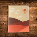 Modern greeting card featuring a warm illustrated landscape with a bright sun and the message "You Brighten My Day" on a soft neutral background.