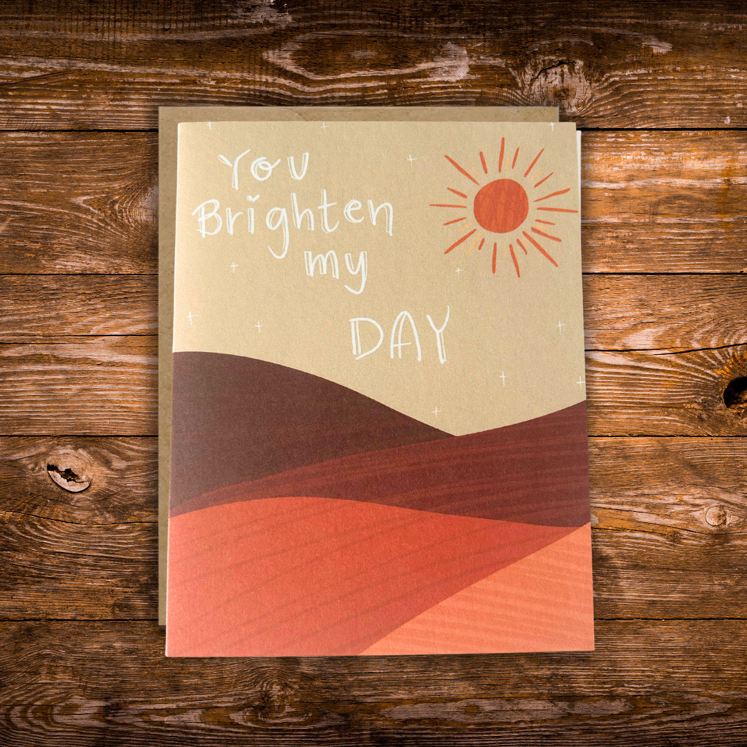 Modern greeting card featuring a warm illustrated landscape with a bright sun and the message "You Brighten My Day" on a soft neutral background.