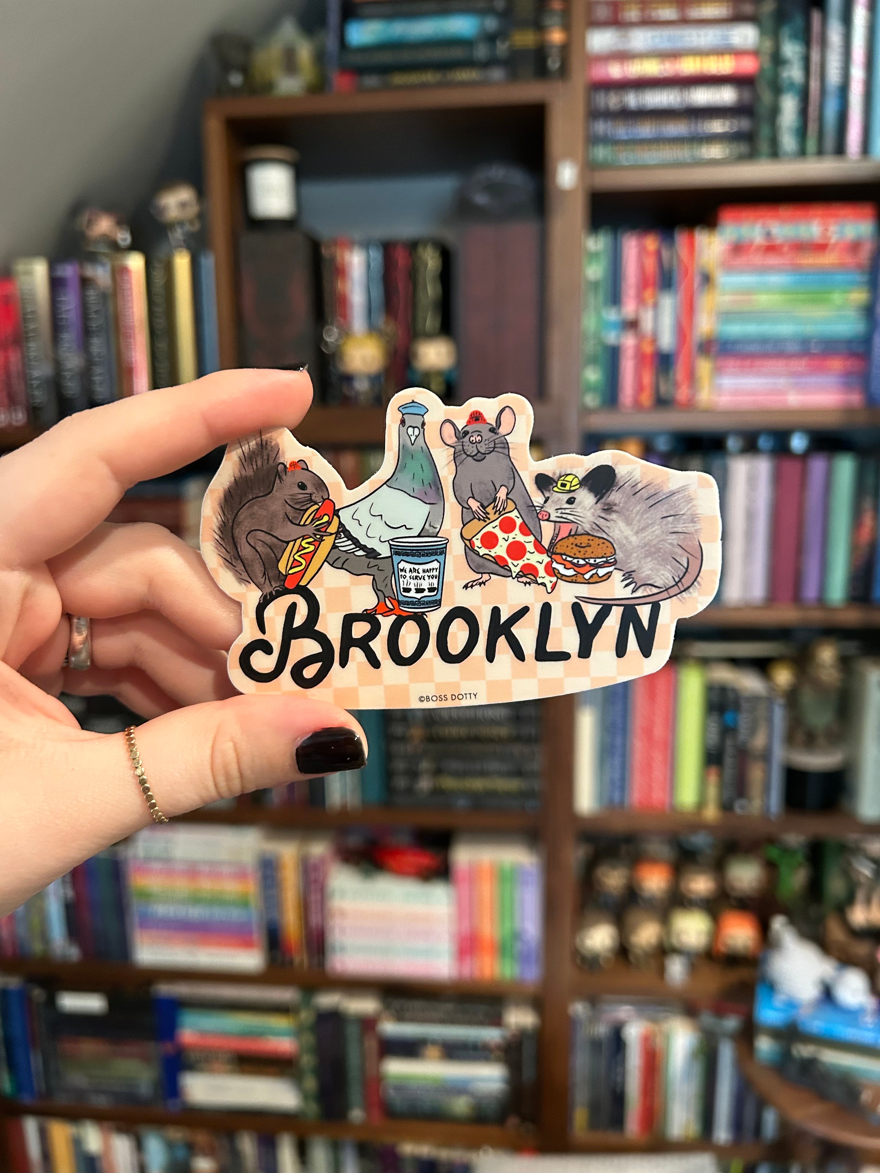 A hand holds a die-cut sticker featuring cartoon animals and the word 'BROOKLYN' against a blurred background of bookshelves.