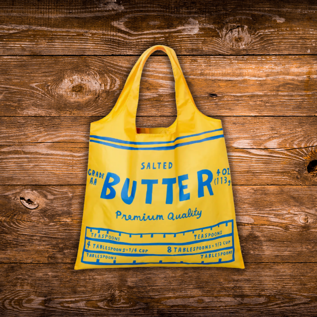 A bright yellow reusable shopping bag styled like a vintage butter milk carton, featuring blue text and measurement lines, displayed on a rustic dark wood background.