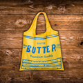 A bright yellow reusable shopping bag styled like a vintage butter milk carton, featuring blue text and measurement lines, displayed on a rustic dark wood background.