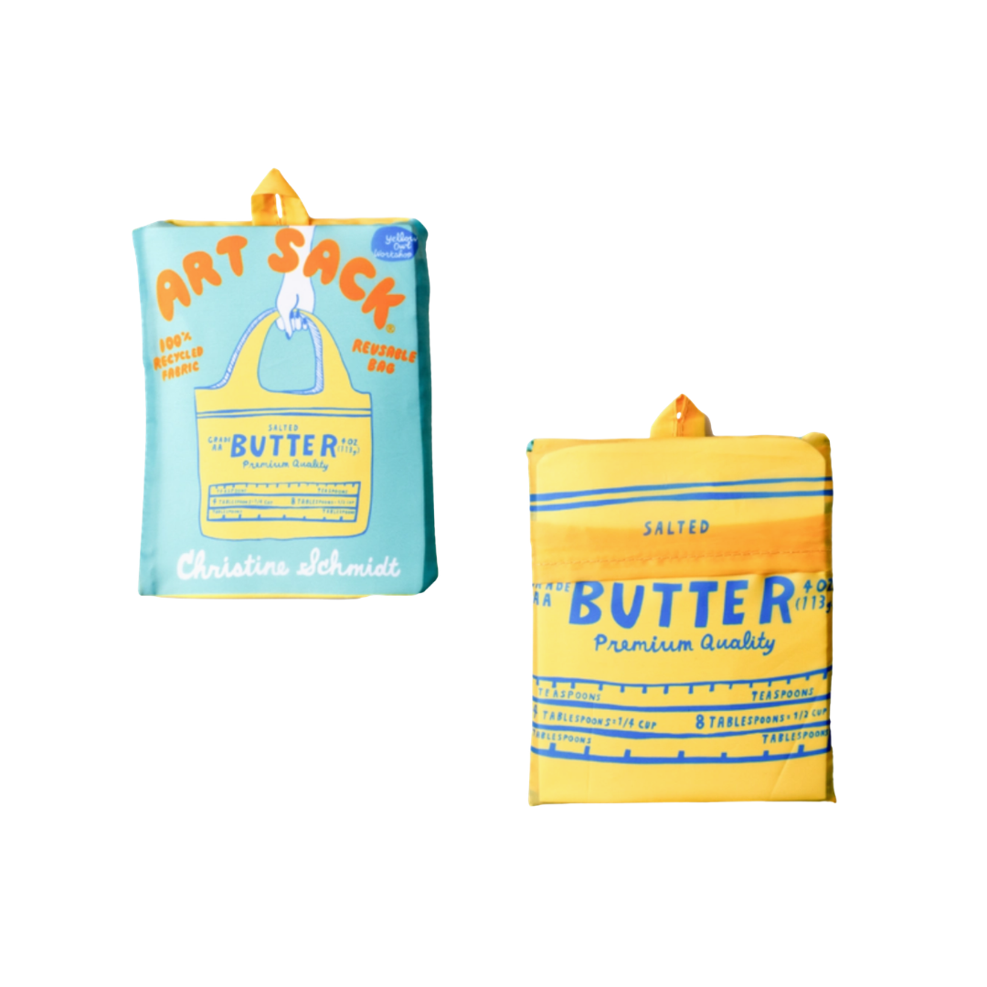 Reusable tote bag shown folded, with the front and back sides visible. The front features a light turquoise background with orange “ART SACK” lettering, a yellow butter-package illustration, and the name “Christine Schmidt.” The back features a close-up yellow butter-package design with blue lettering, measuring marks, and orange accents.