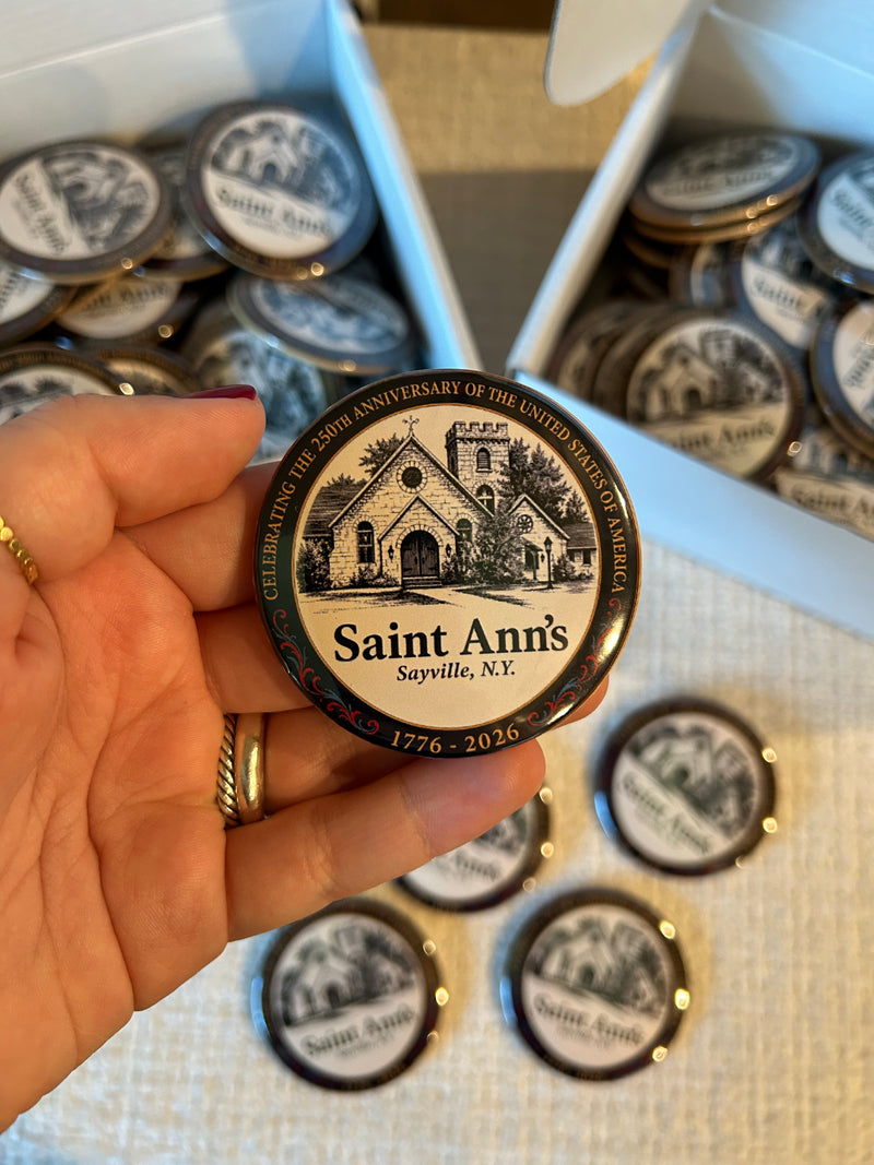 A hand holds a round Saint Ann's church commemorative pin badge in front of open boxes filled with similar badges.