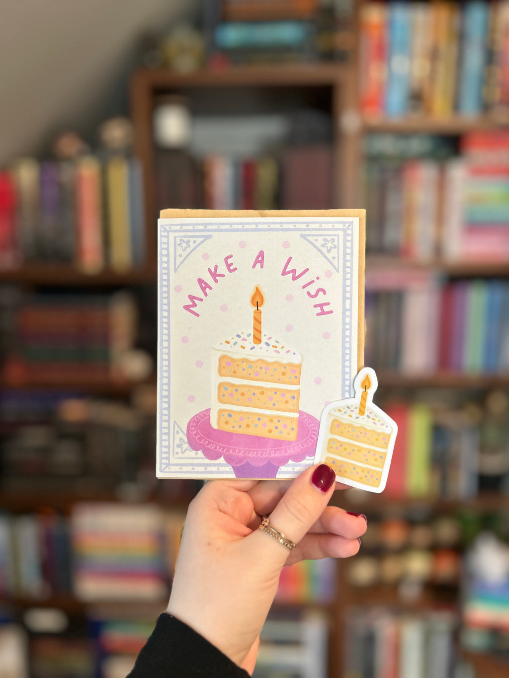 Matching birthday greeting card and waterproof vinyl sticker featuring a pastel slice of birthday cake with sprinkles, a lit candle, and the words "Make a Wish."