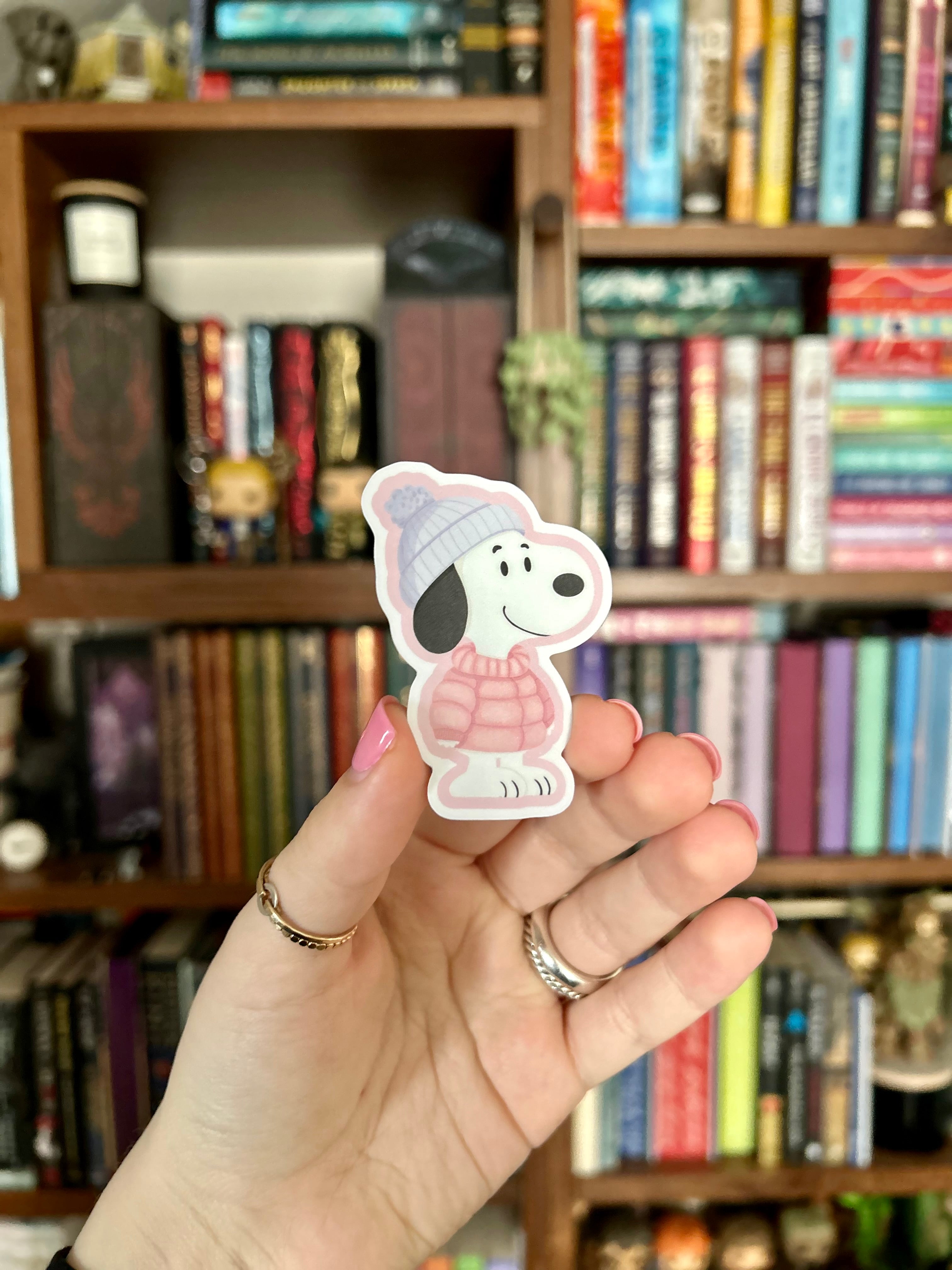 A hand holding a die-cut sticker of a Beagle wearing a pink sweater and gray beanie, with a blurred bookshelf in the background.