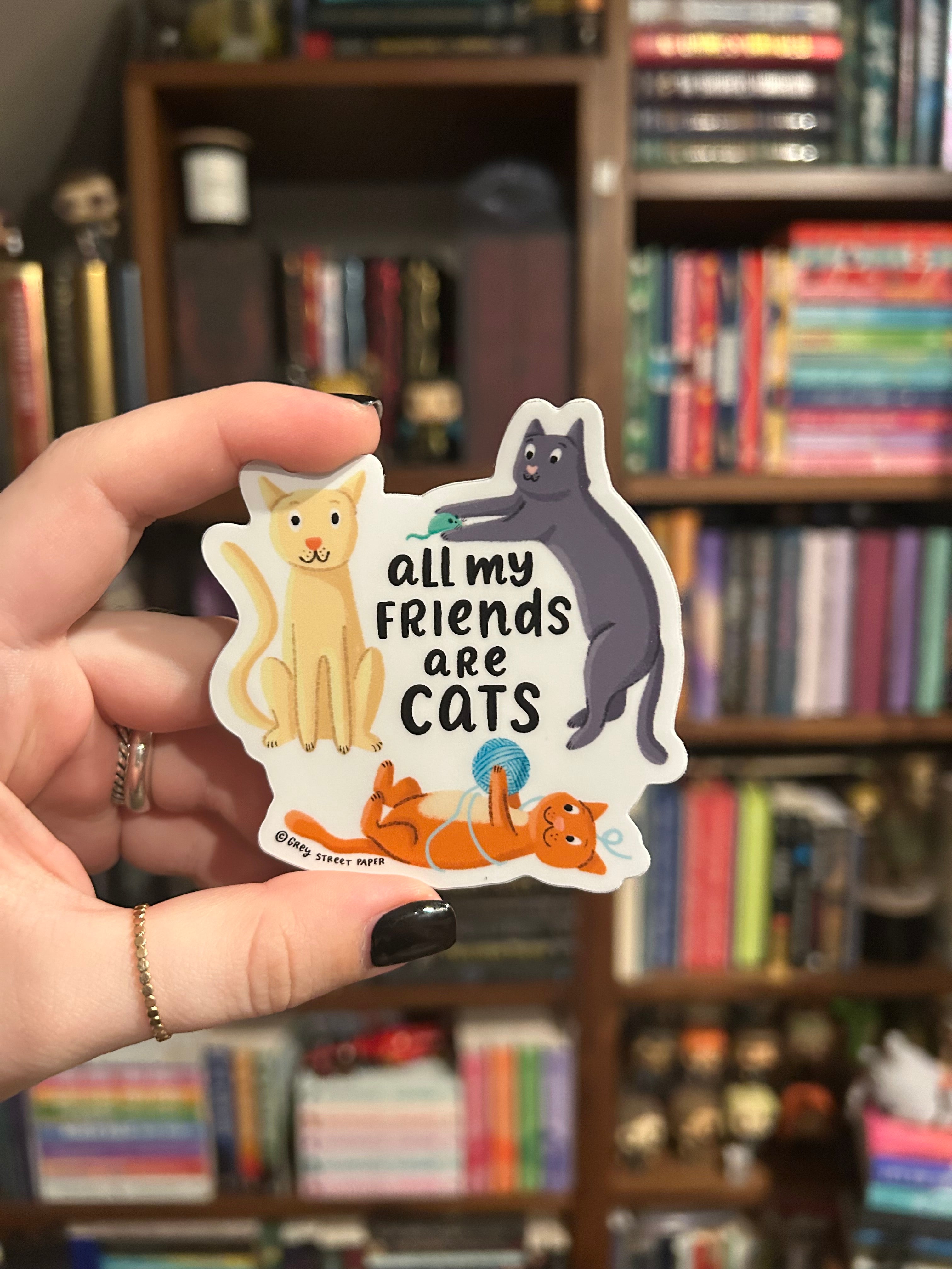 A hand with black nail polish holds a die-cut sticker featuring cartoon cats and the text 'all my Friends are CATS' in front of a blurred bookshelf background.