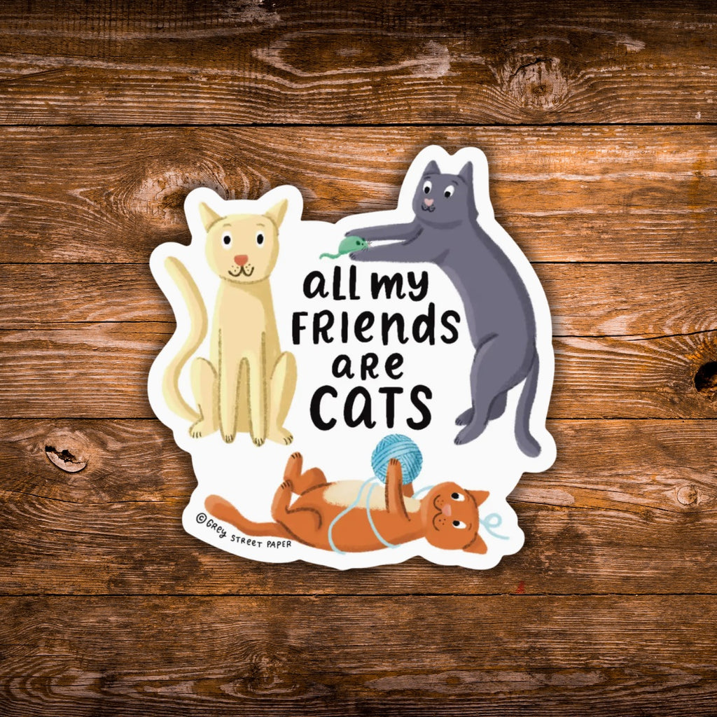 A die-cut sticker featuring three cartoon cats and the text 'all my FRIENDS are CATS' on a white background, placed on a rustic brown wood plank surface. The sticker includes a cream cat on the left, a dark gray cat on the right pushing a green toy, and an orange cat lying on its back below.