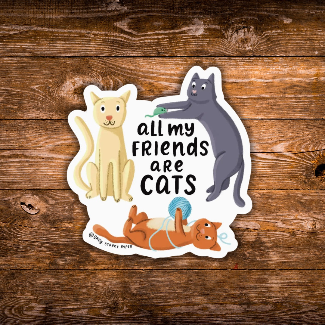 A die-cut sticker featuring three cartoon cats and the text 'all my FRIENDS are CATS' on a white background, placed on a rustic brown wood plank surface. The sticker includes a cream cat on the left, a dark gray cat on the right pushing a green toy, and an orange cat lying on its back below.