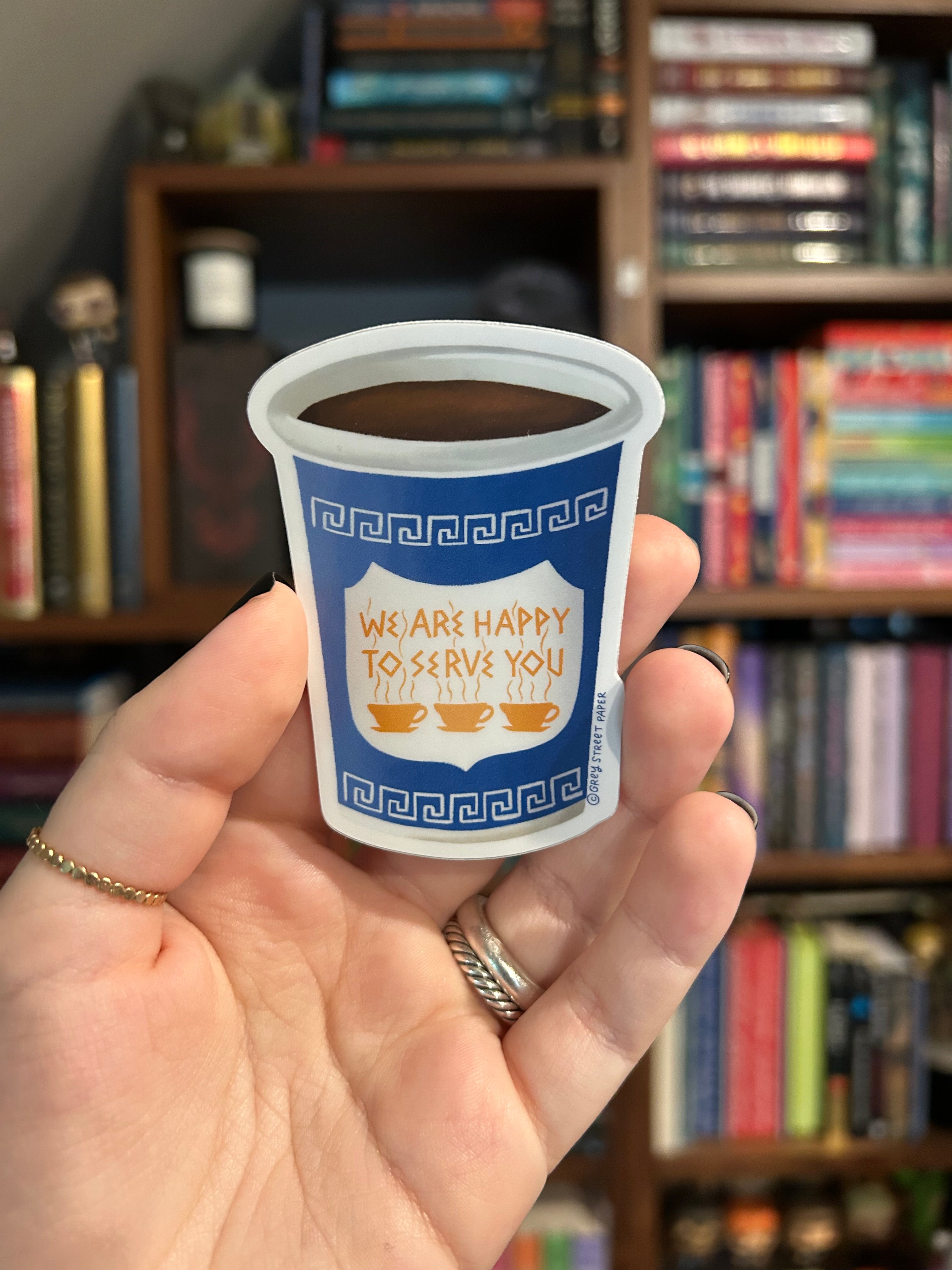 A hand holding a sticker of a white coffee cup with a blue label that reads 'WE ARE HAPPY TO SERVE YOU' and shows a blurred bookshelf in the background.
