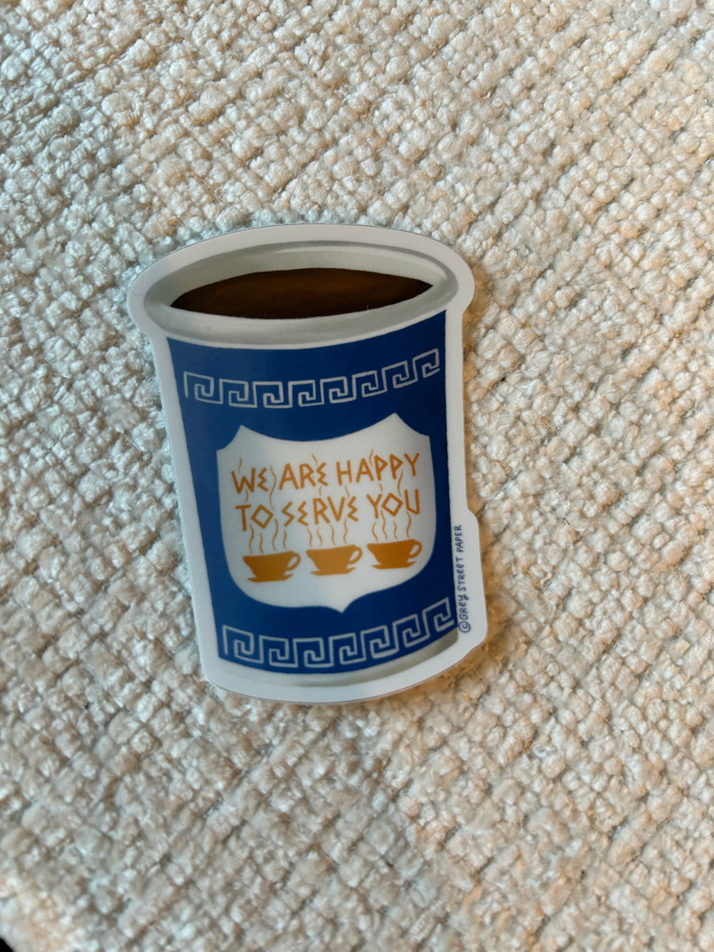 A sticker of a coffee cup with a Greek key borders, three coffee cups, and the phrase 'WE ARE HAPPY TO SERVE YOU'.