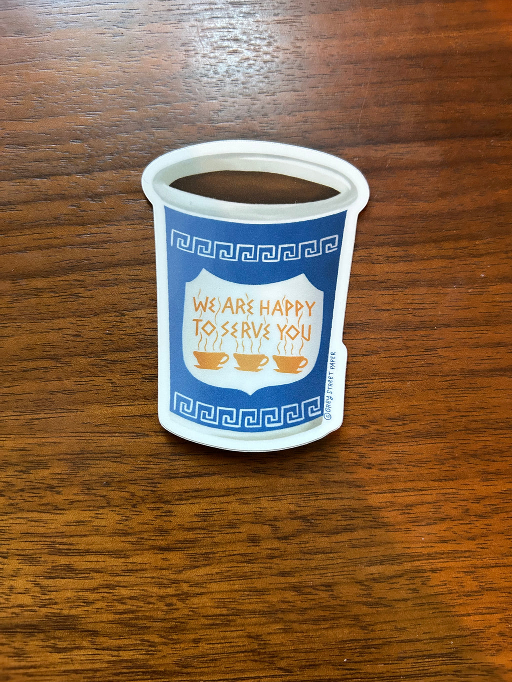 A small white coffee cup sticker with a blue and white printed design featuring the text 'WE ARE HAPPY TO SERVE YOU' and coffee cup illustrations, placed on a brown wooden surface.