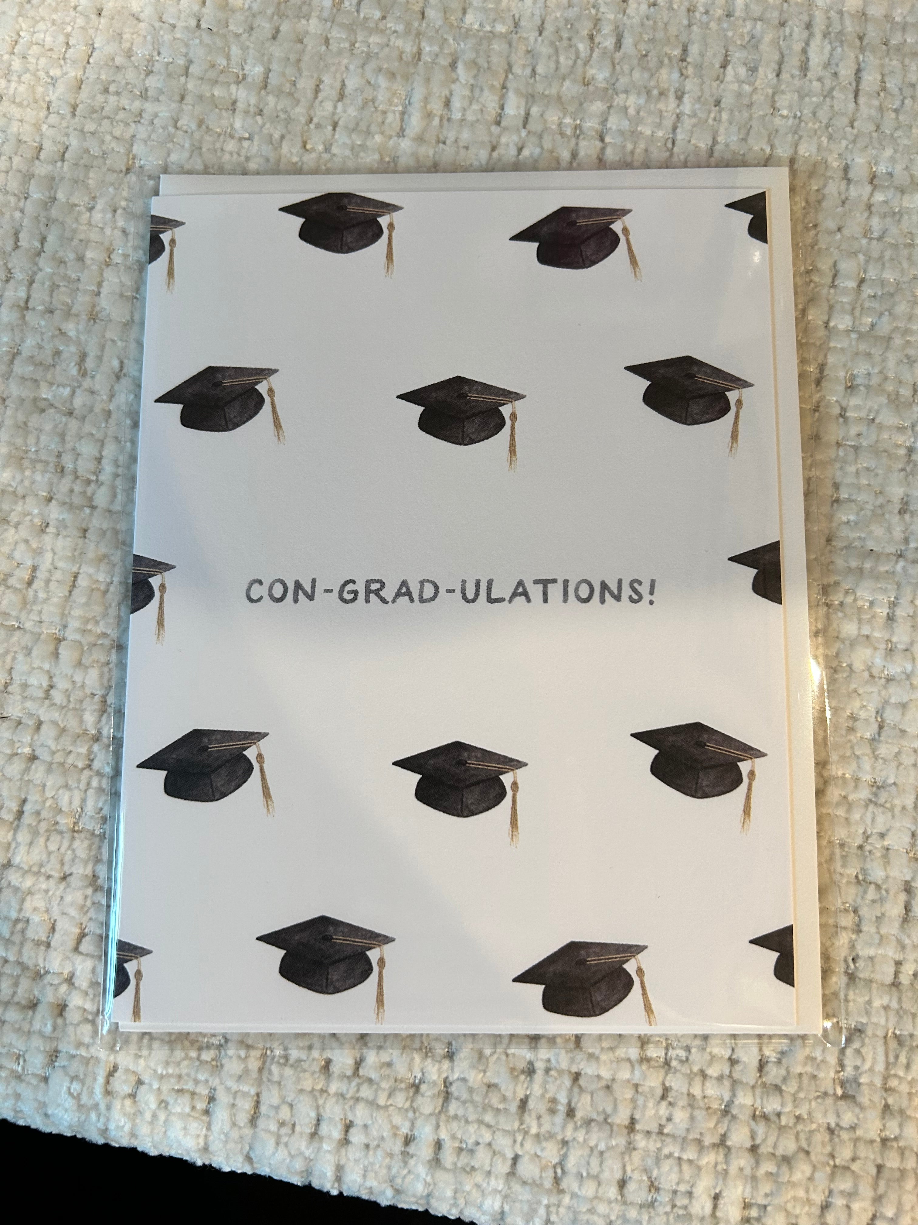 Graduation greeting card featuring a repeating pattern of graduation caps with the pun "Con-Grad-Ulations!" centered on a white background.