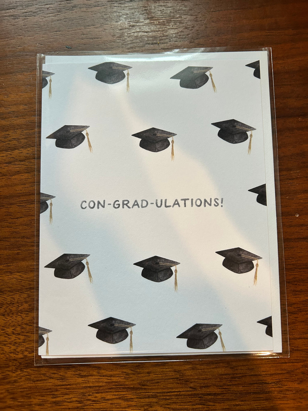 Graduation greeting card featuring a repeating pattern of graduation caps with the pun "Con-Grad-Ulations!" centered on a white background.