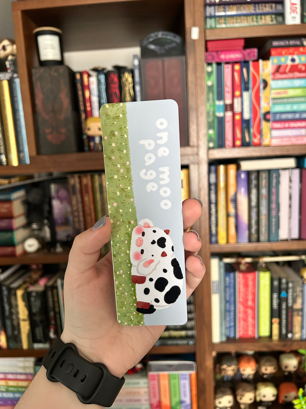 Double-sided cow bookmark featuring a rosy-cheeked white cow in a green grassy field with playful lettering, paired with a black-and-white cow print reverse.