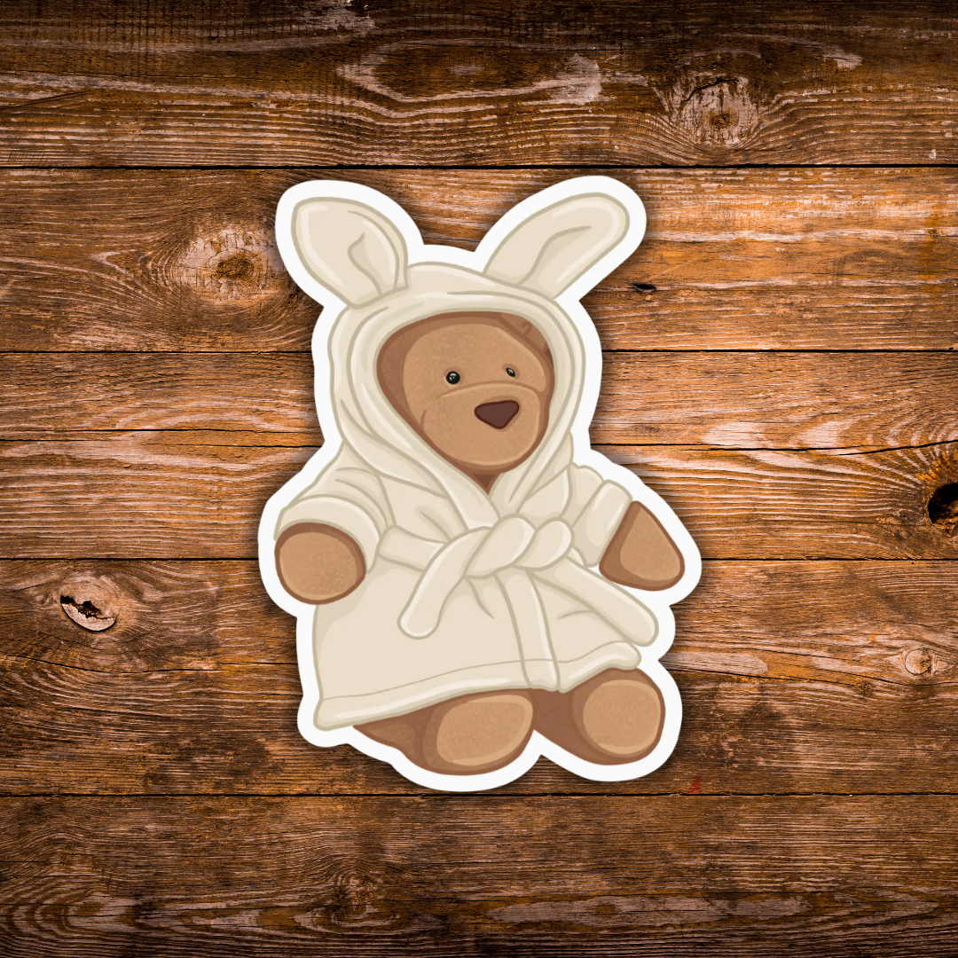 A die-cut sticker featuring a cartoon teddy bear wearing a white bunny costume, placed on a rustic brown wooden plank background.