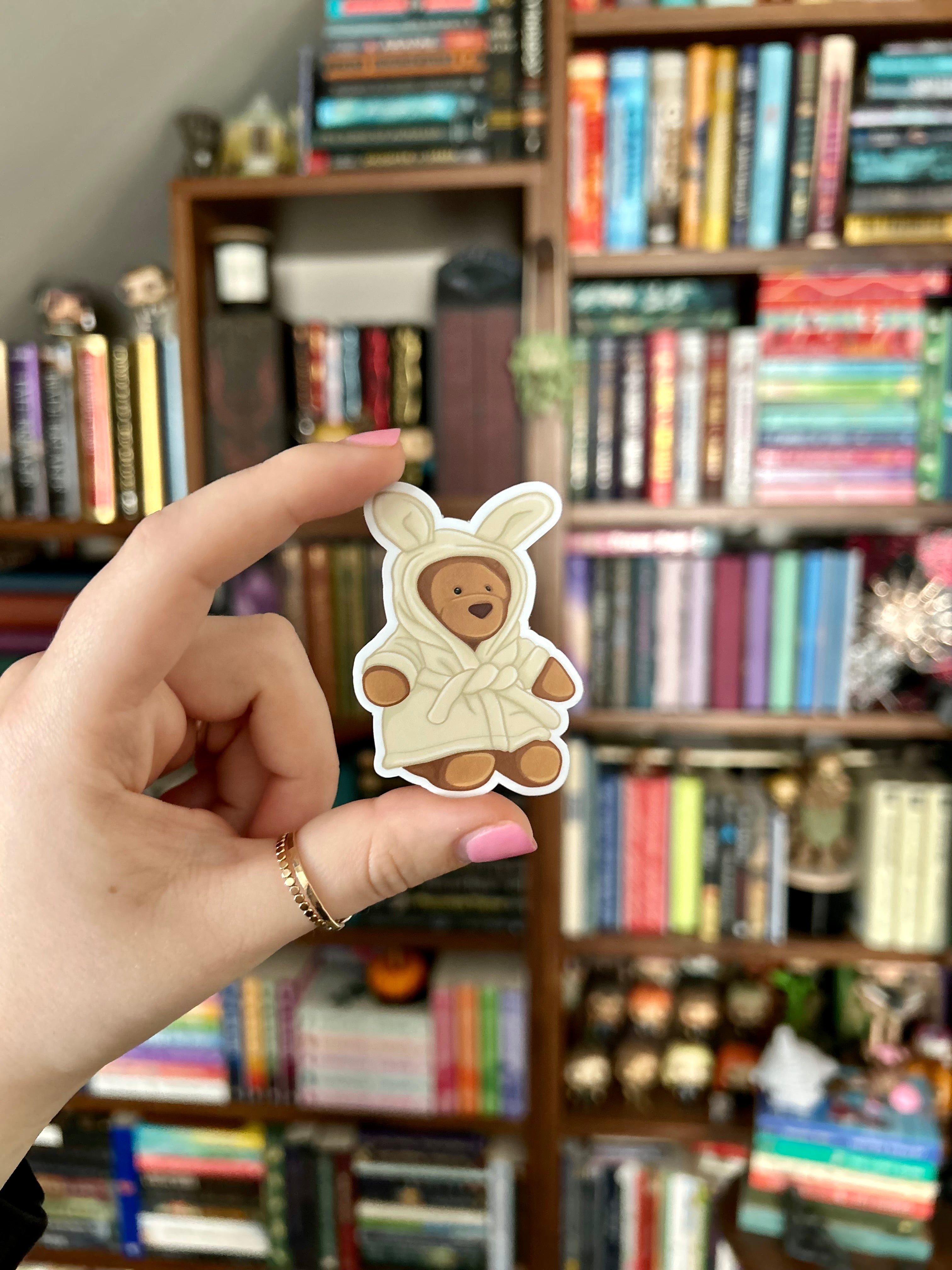 A hand holds a small die-cut sticker or patch featuring a teddy bear in a cream bunny costume, with a blurred bookshelf background.