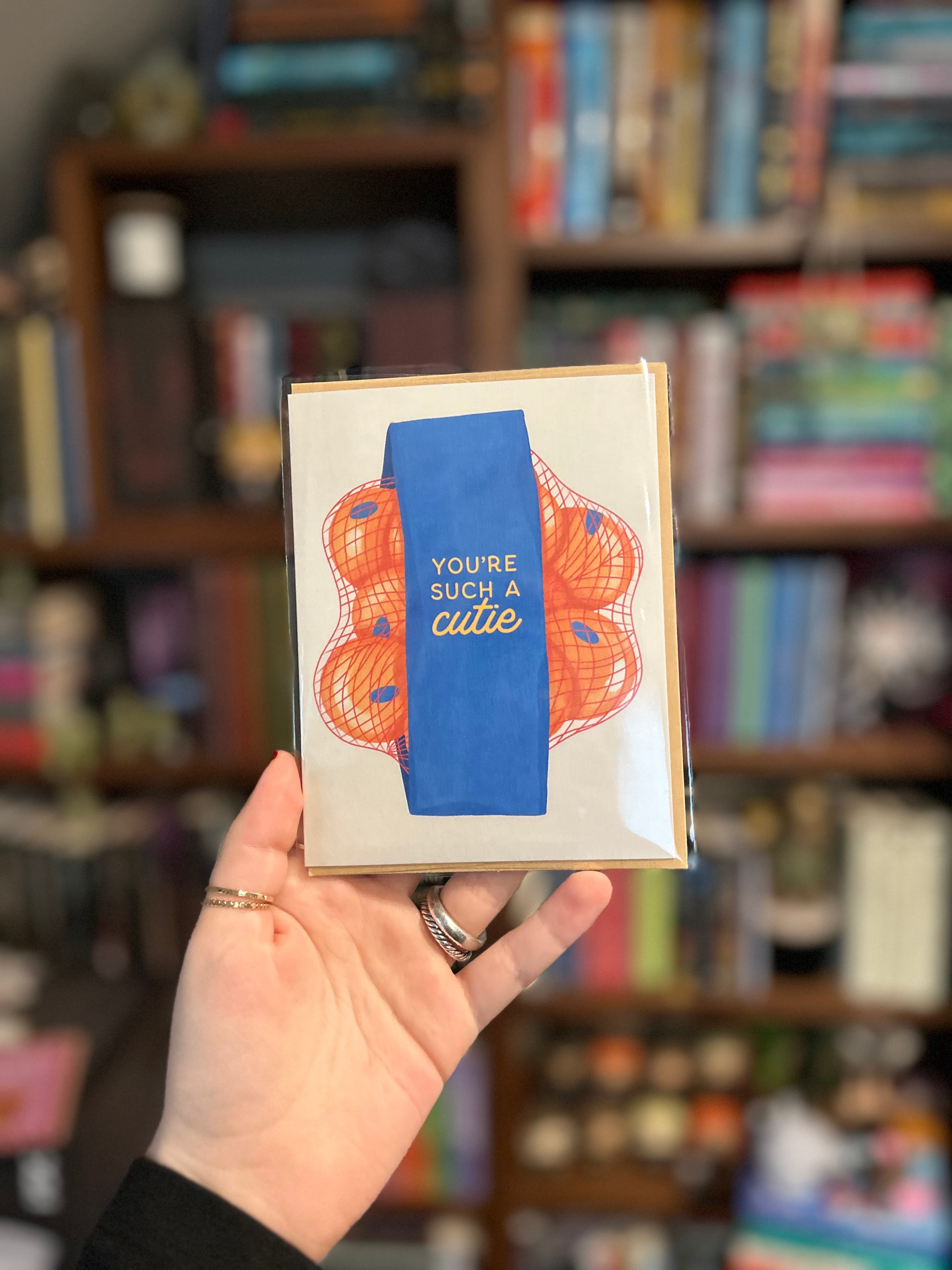 Greeting card featuring a bag of clementines with the phrase "You're Such a Cutie" on a blue label against a neutral background.
