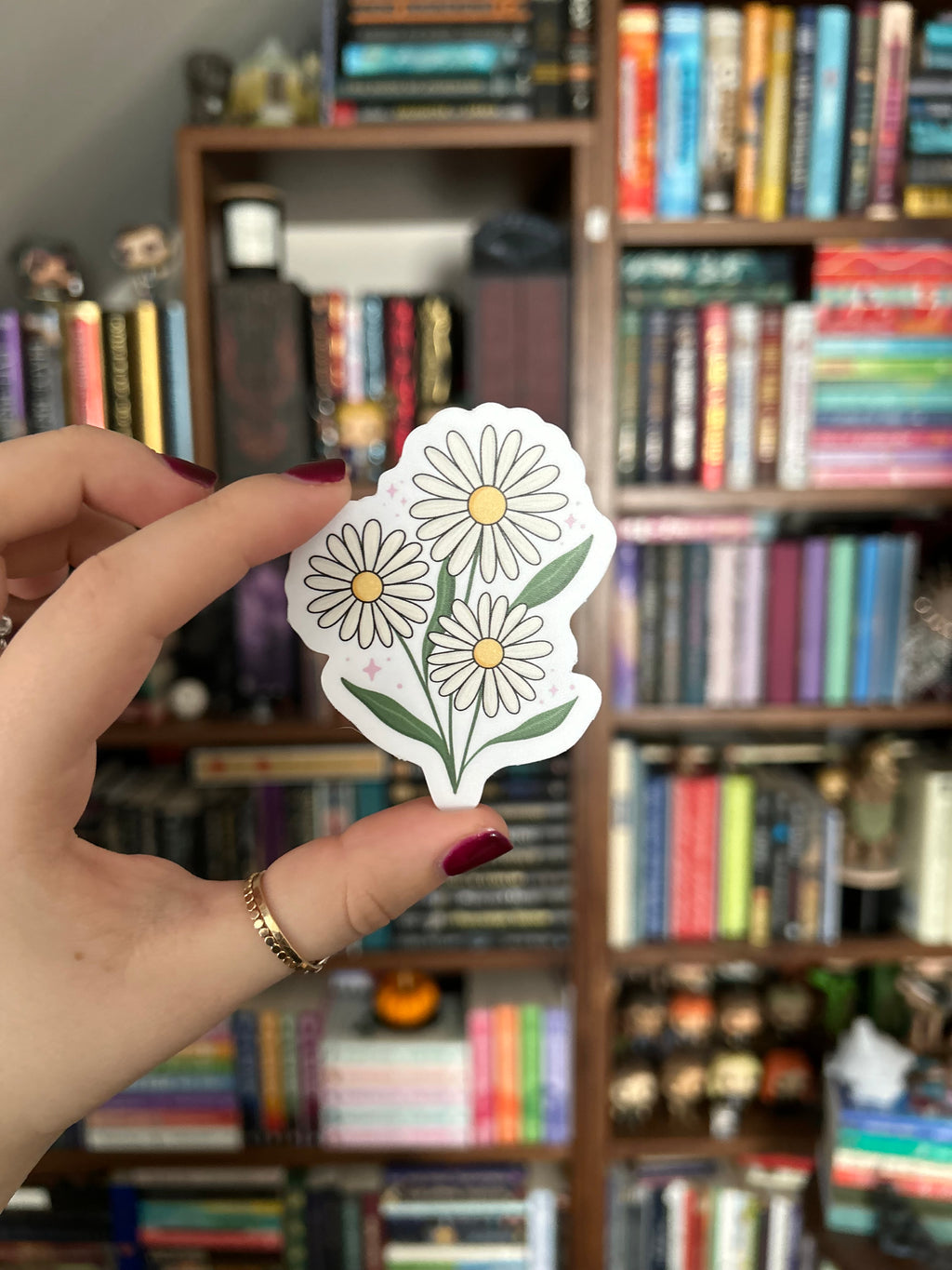 A hand with dark red nail polish holds a die-cut sticker featuring three white daisy flowers with yellow centers and green leaves, set against a blurred background of bookshelves.