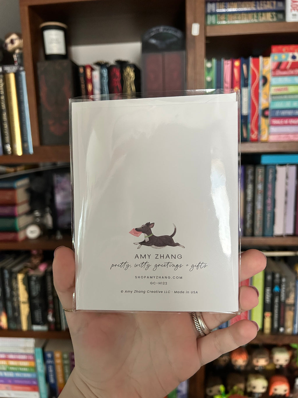 A hand holding a clear plastic sleeve containing a white greeting card with a black dog illustration and the text 'AMY ZHANG pretty witty greetings gifts' in front of a bookshelf filled with books.