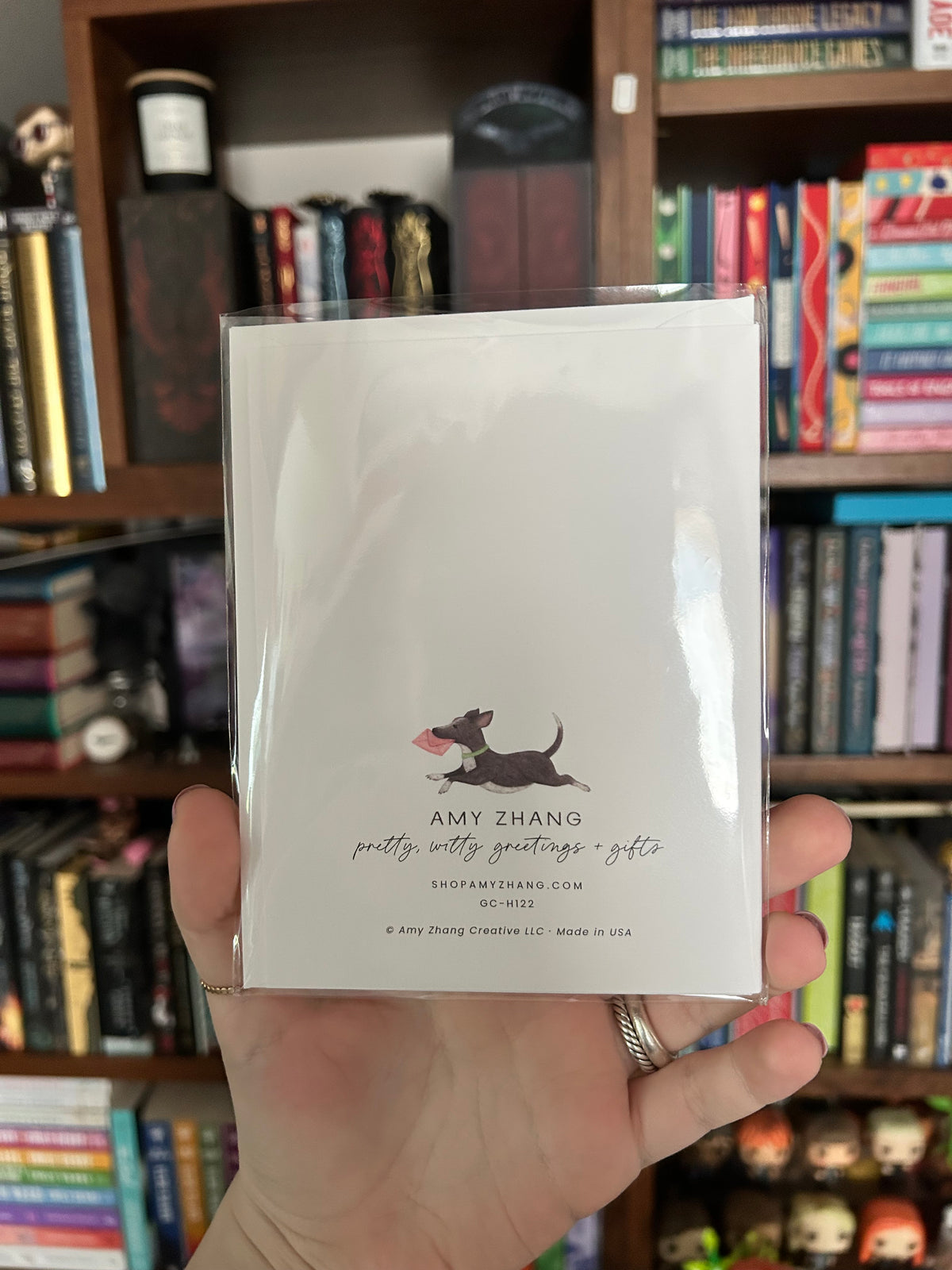 A hand holding a clear plastic sleeve containing a white greeting card with a black dog illustration and the text 'AMY ZHANG pretty witty greetings gifts' in front of a bookshelf filled with books.