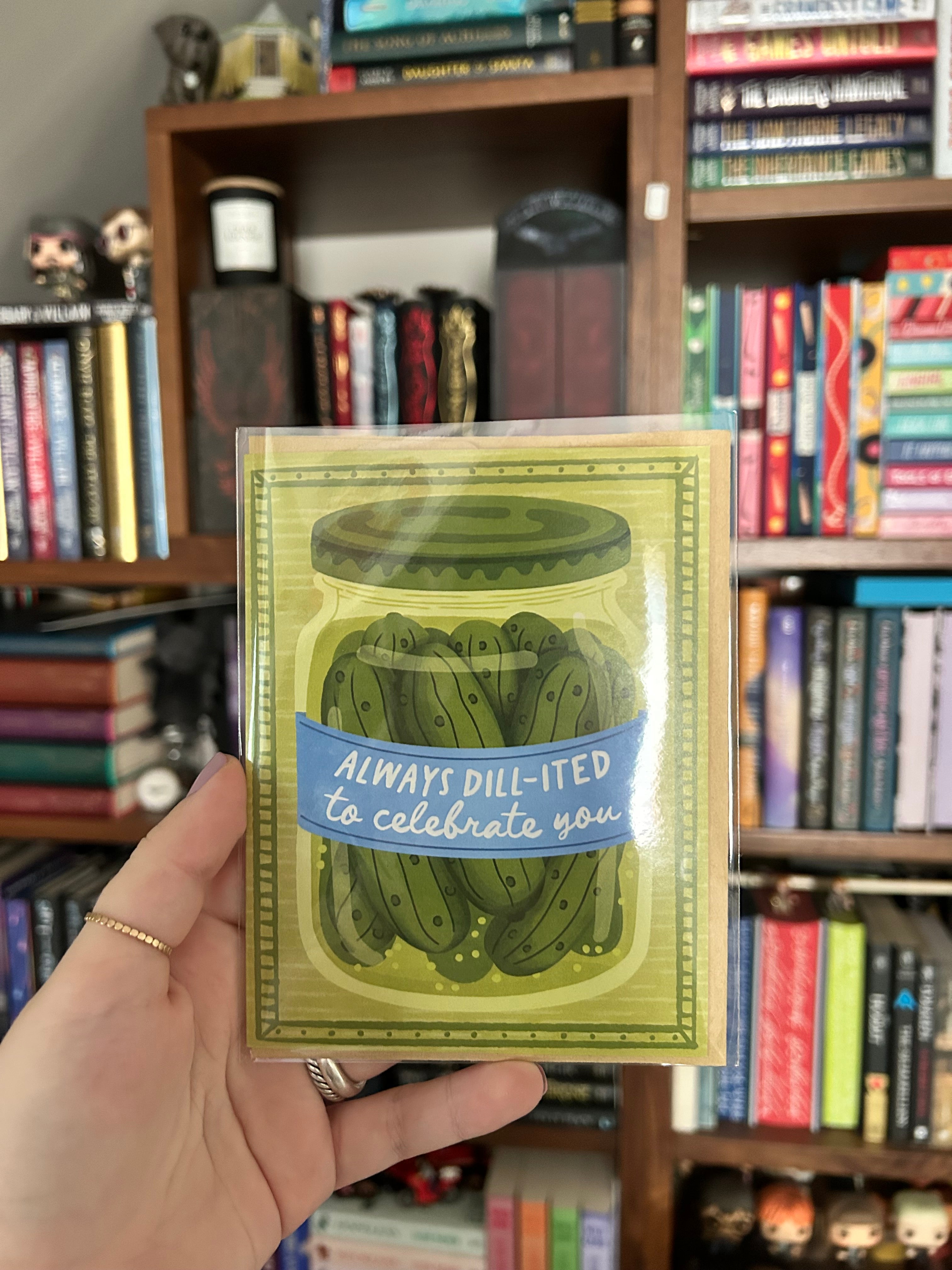 A hand holds a greeting card featuring an illustration of pickled dill greens in a glass jar with the text 'ALWAYS DILL-ITED to celebrate you'. The background shows a bookshelf filled with books.