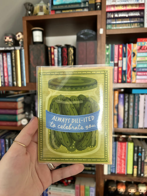 A hand holds a greeting card featuring an illustration of pickled dill greens in a glass jar with the text 'ALWAYS DILL-ITED to celebrate you'. The background shows a bookshelf filled with books.