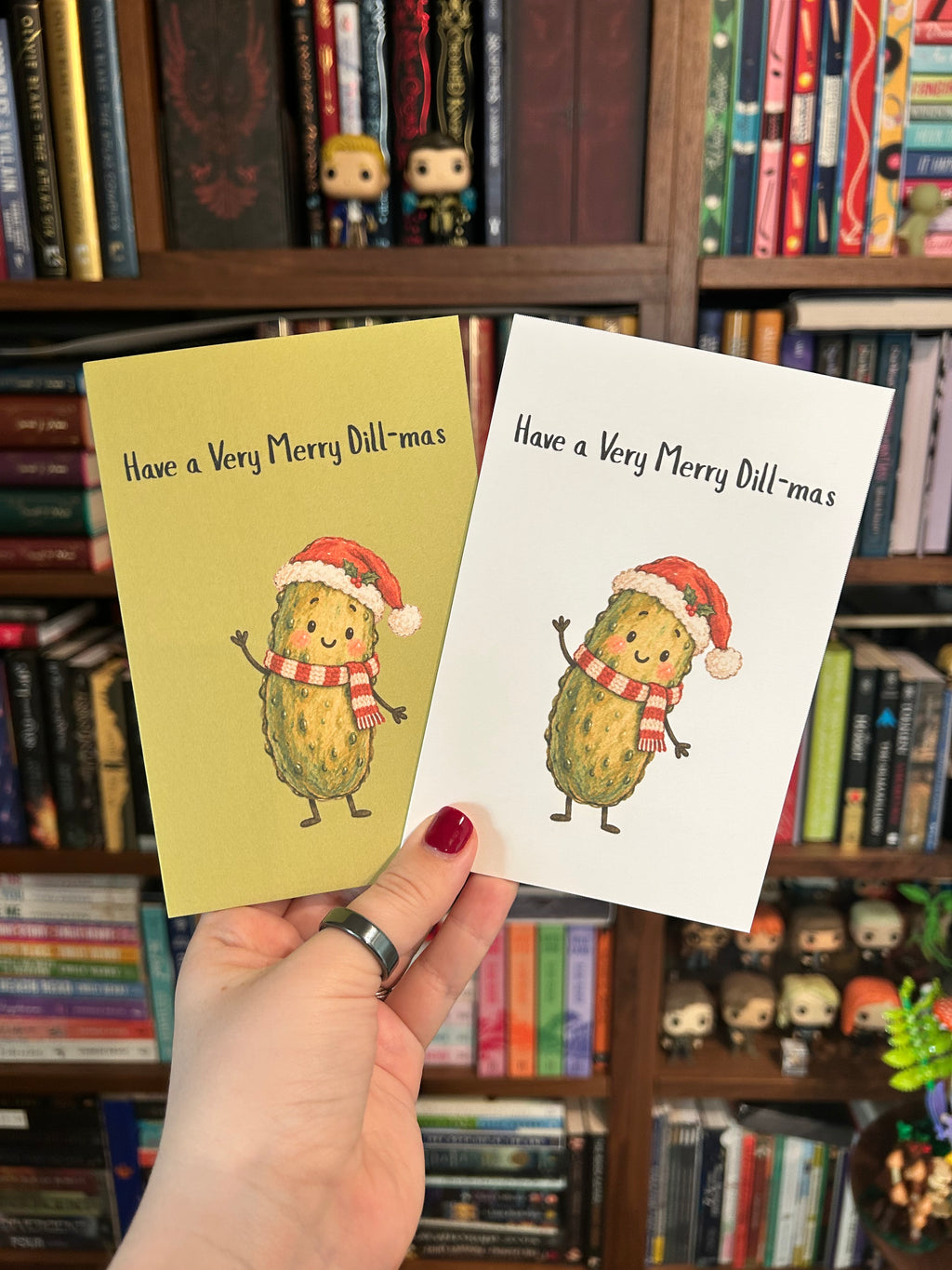 Christmas greeting card featuring a smiling green pickle wearing a Santa hat and red-and-white scarf with the phrase “Have a Very Merry Dill-mas.” Available with a white or olive-green background and a kraft envelope.