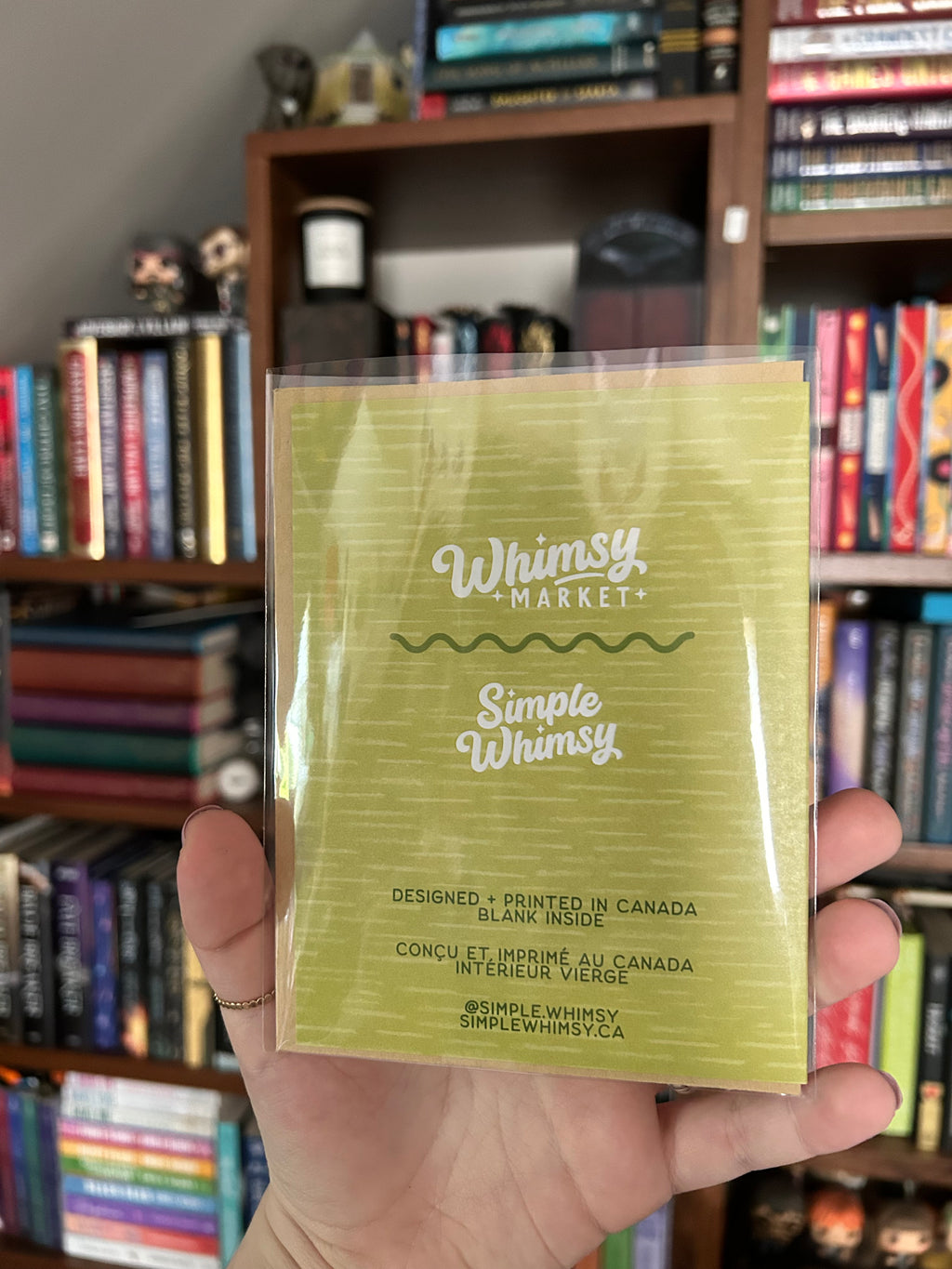 A hand holds a clear plastic-wrapped card of Whimsy Market Simple Whimsy card in front of a blurred bookshelf background.