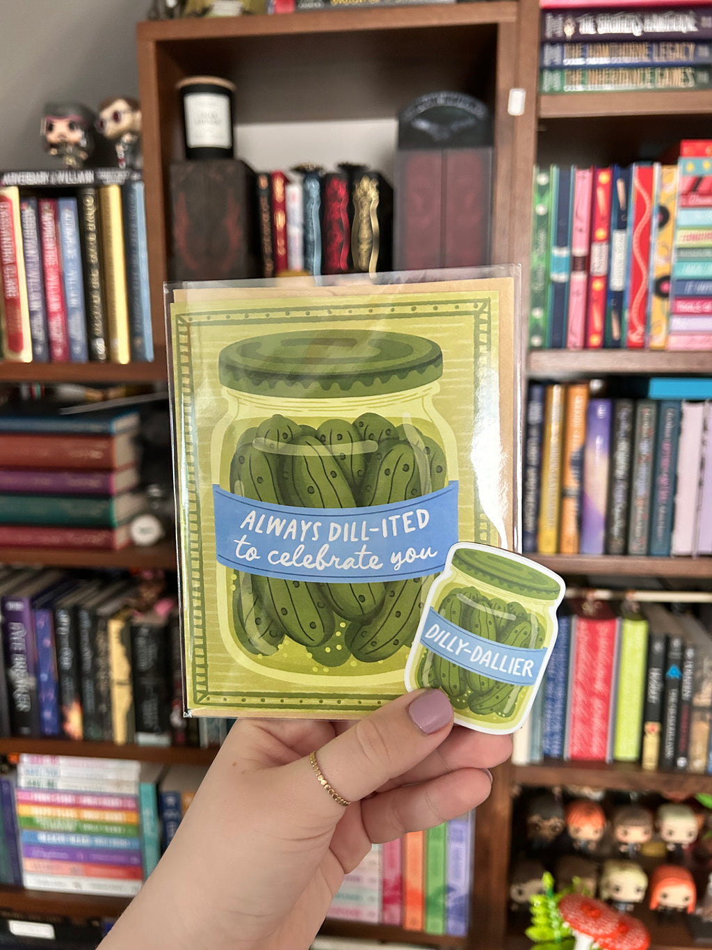 A hand holds a packaged greeting card and matching sticker featuring an illustrated pickle jar with the text 'ALWAYS DILLY-ITED to celebrate you' and 'DILLY-DALLIER'. The background shows a real indoor environment with bookshelves filled with books.