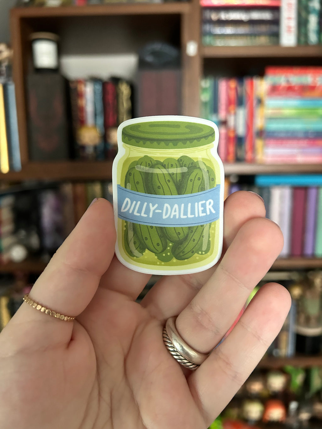 A hand holding a die-cut sticker of a pickle jar labeled 'DILLY-DALLIER' against a blurred background of bookshelves.