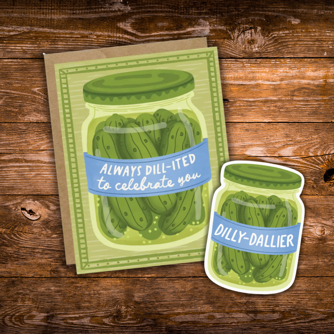 A greeting card and matching die-cut sticker featuring an illustrated jar of pickles with a blue banner reading 'ALWAYS DILL-ITED to celebrate you' and 'DILLY-DALLIER', displayed on a rustic wooden surface.