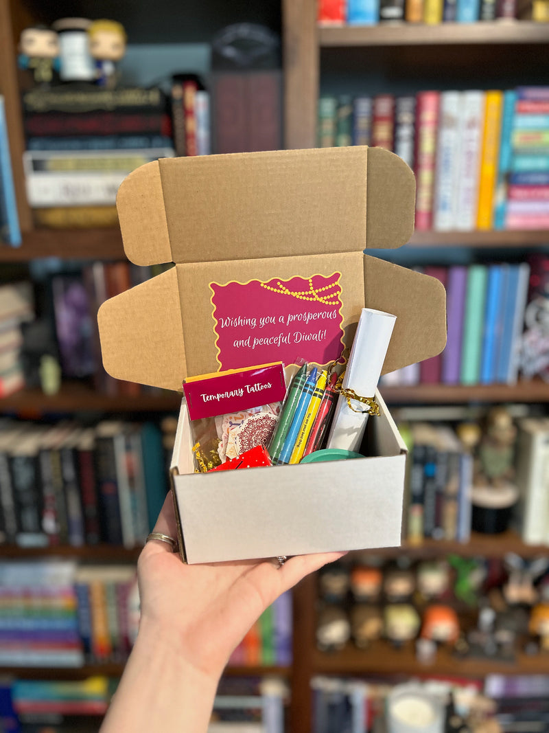 A hand holds an open cardboard gift box filled with assorted items, including colorful crayon, coloring pages, and a temporary tattoos, in front of a blurred bookshelf background.