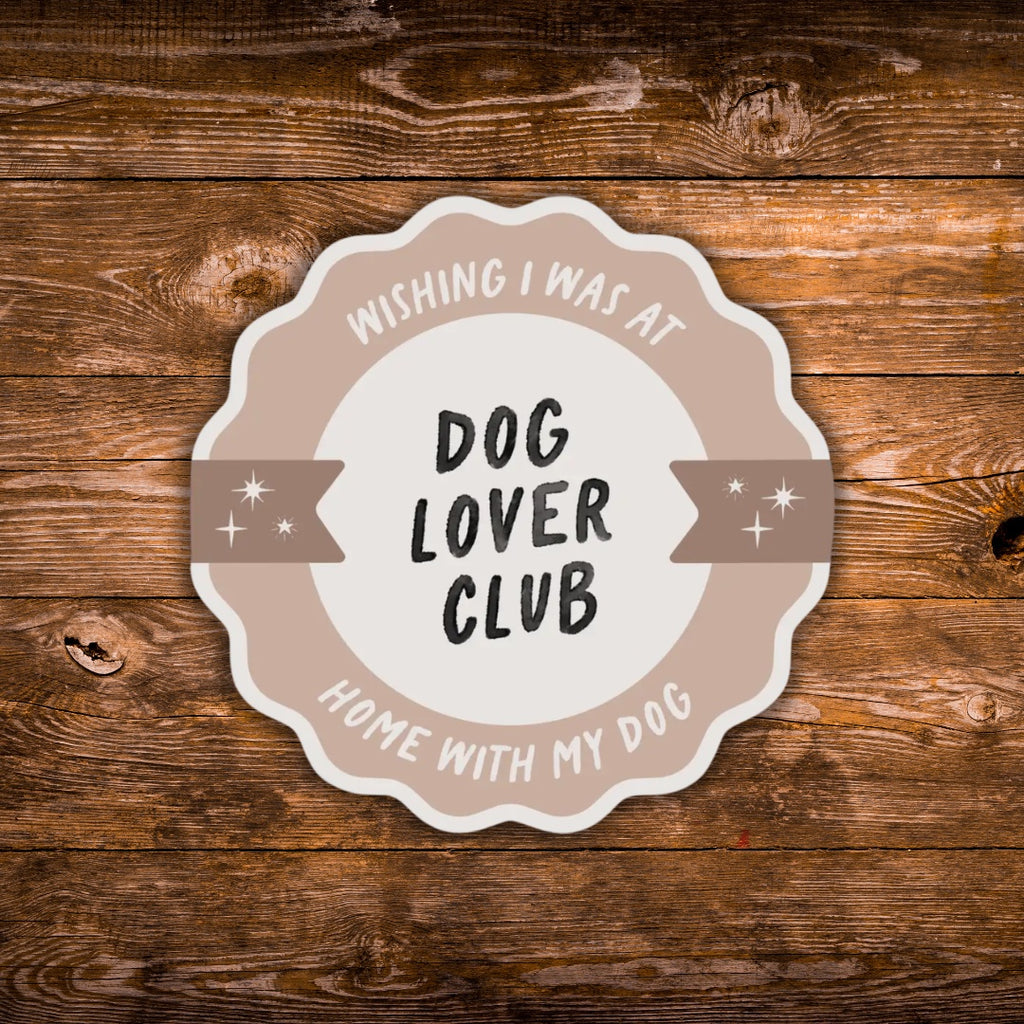 A scalloped circular sticker or decal with the text 'WISHING I WAS AT HOME WITH MY DOG- DOG LOVER CLUB' in black and beige lettering, shown on a rustic brown wood plank background.