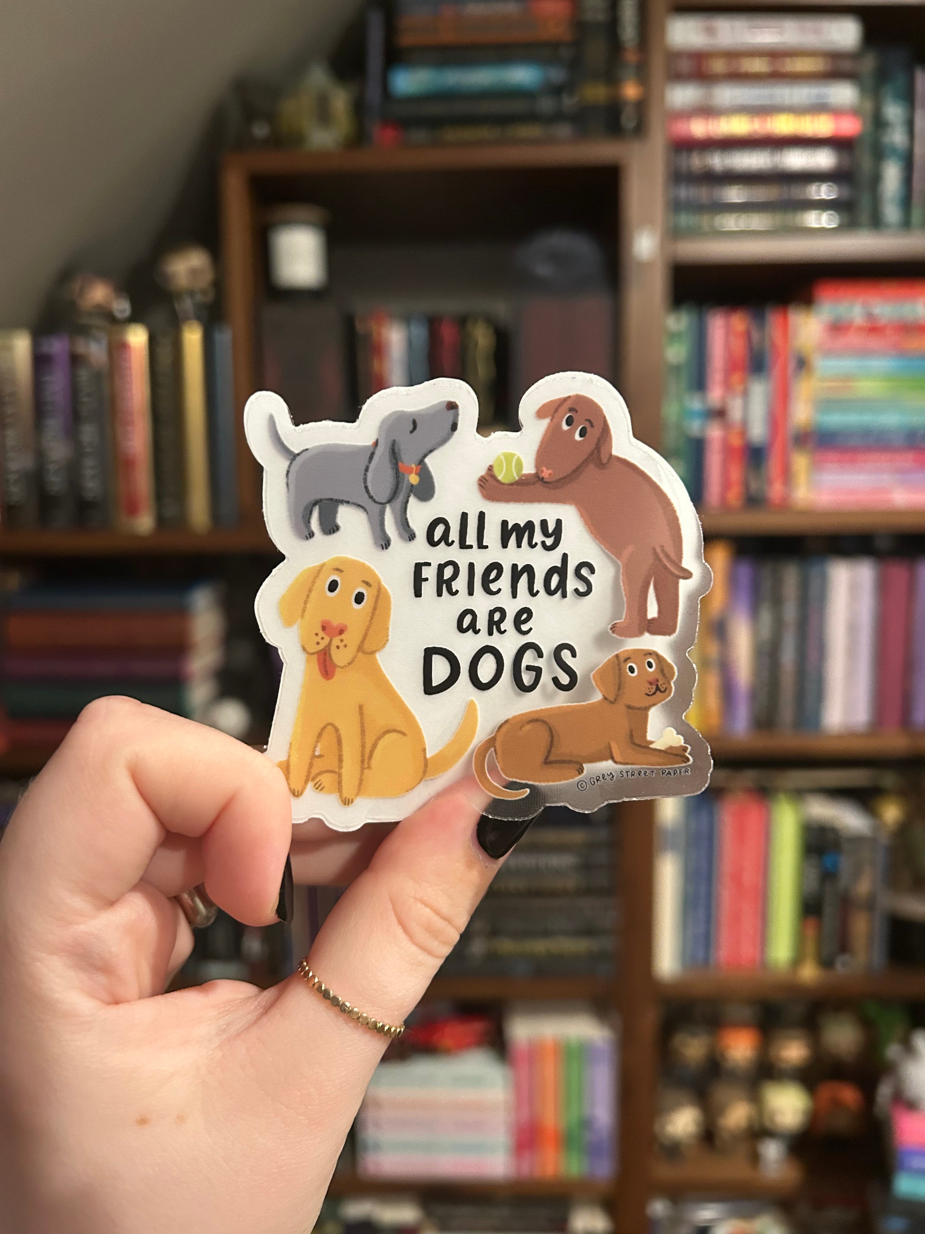 A hand holds a die-cut sticker featuring cartoon dogs and the text 'all my friends are DOGS' against a blurred bookshelf background.