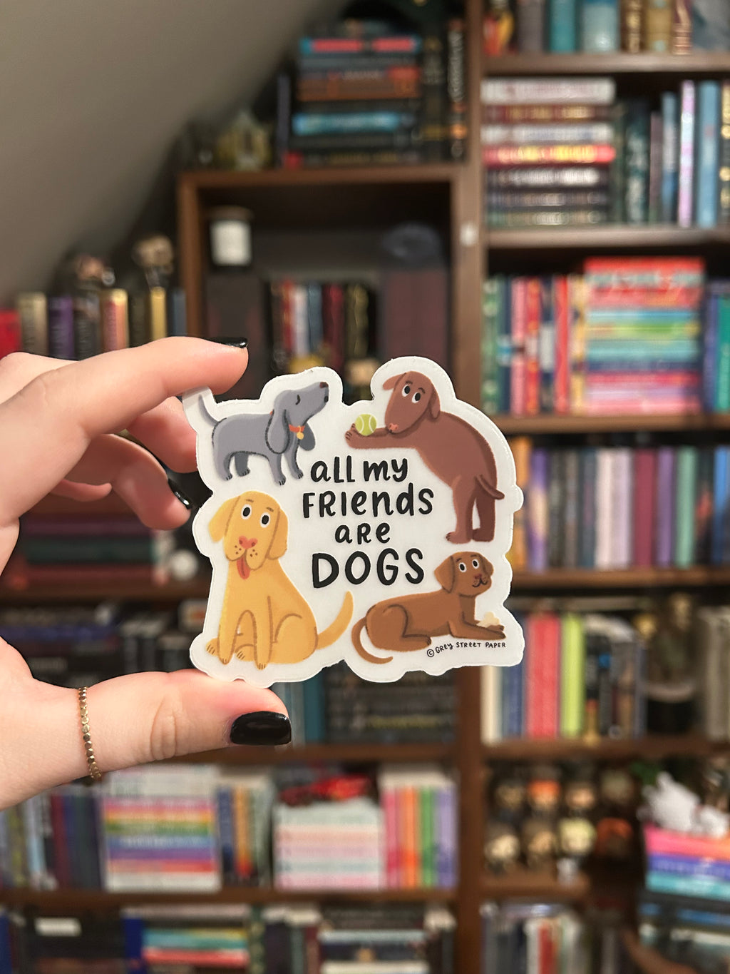 A hand holds a die-cut sticker featuring cartoon dogs and the text 'all my friends are DOGS' in front of a blurred bookshelf background.