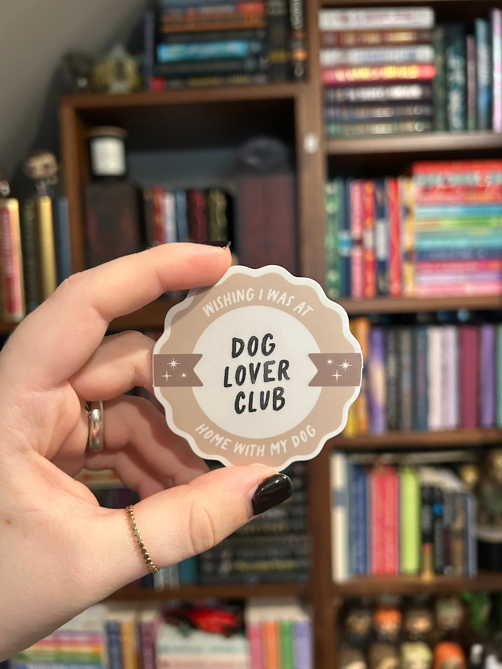 A hand with black nail polish holds a round, scalloped-edge sticker that reads 'WISHING I WAS AT HOME WITH MY DOG- DOG LOVER CLUB' in a blurred bookshelf background.