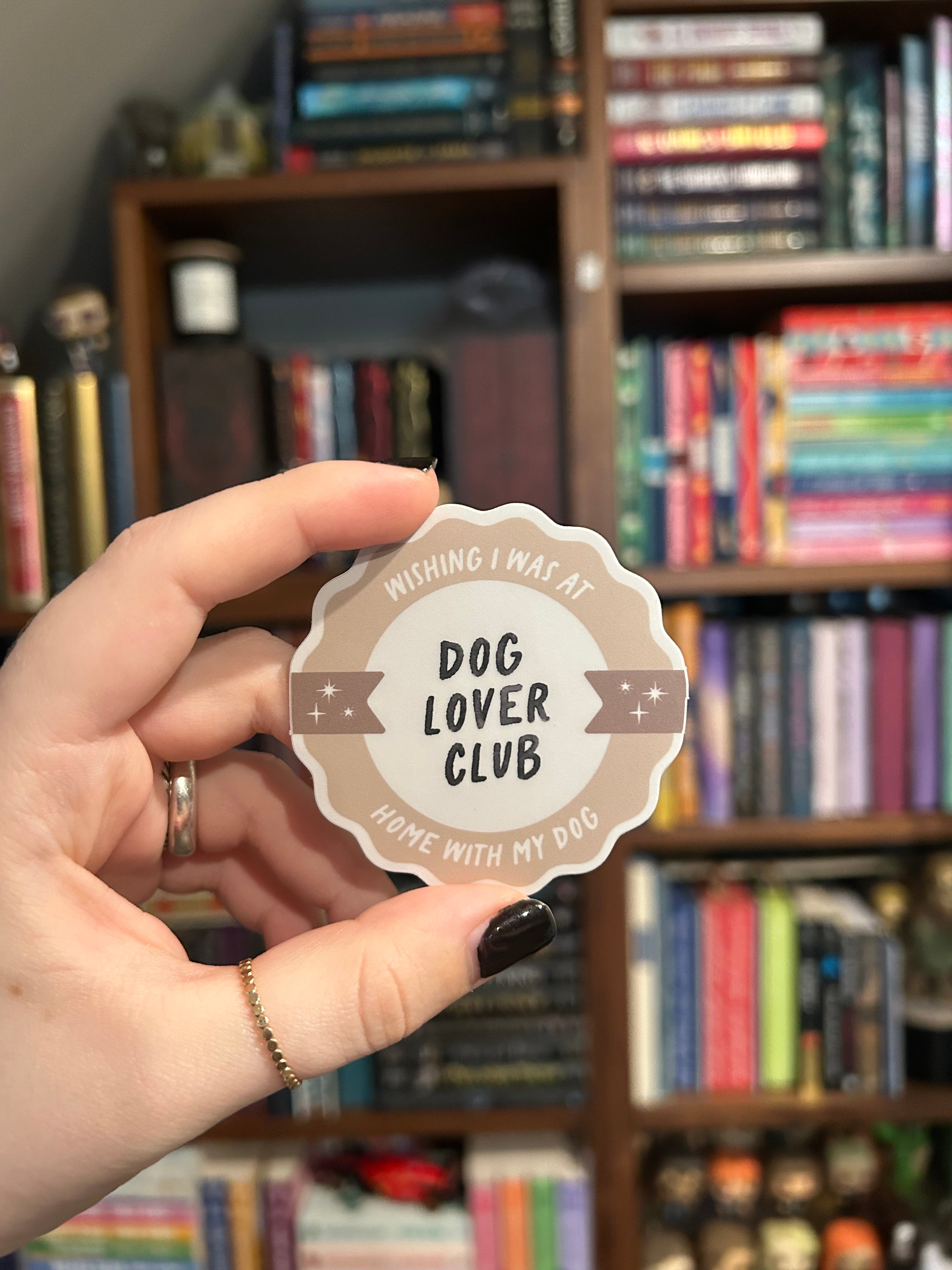 A hand with black nail polish holds a round, scalloped-edge sticker that reads 'WISHING I WAS AT HOME WITH MY DOG- DOG LOVER CLUB' in a blurred bookshelf background.