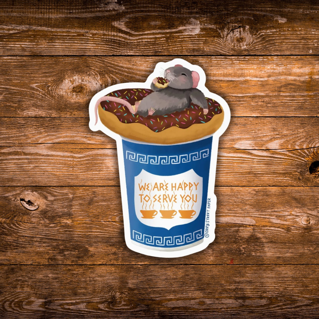 A die-cut sticker featuring a cartoon gray mouse sitting on a donut with sprinkles inside a blue coffee cup, displayed on a rustic brown wooden background. The cup has Greek key patterns and the text 'WE ARE HAPPY TO SERVE YOU'.