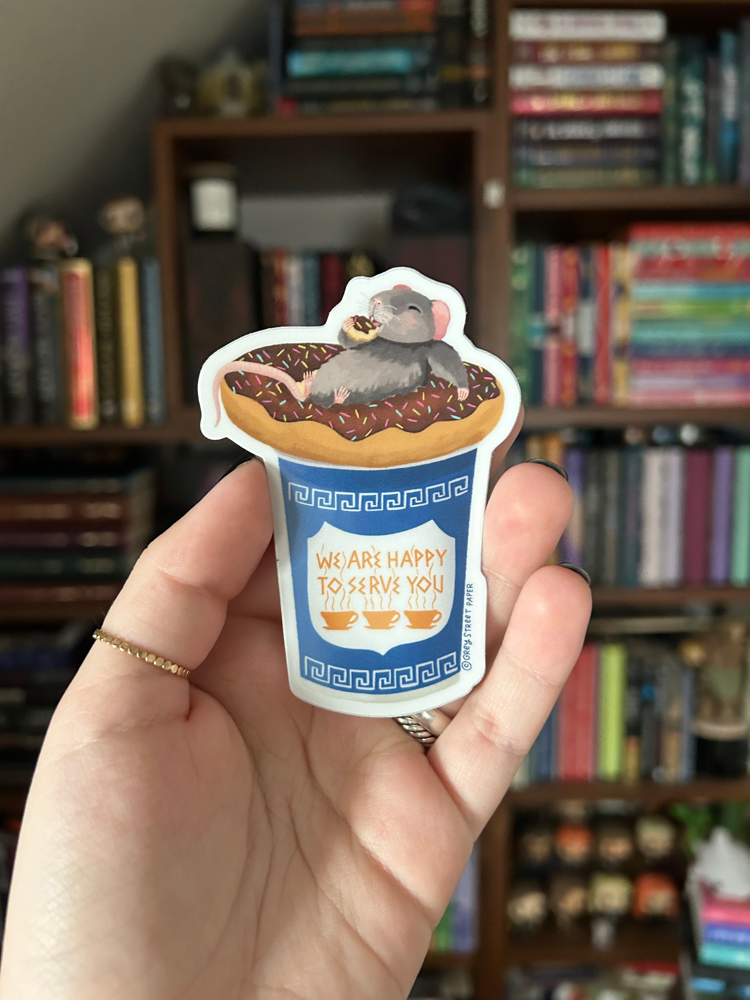 A hand holding a die-cut sticker featuring a gray rat sitting in a coffee cup on a donut with the text 'WE ARE HAPPY TO SERVE YOU', with a blurred bookshelf behind it.