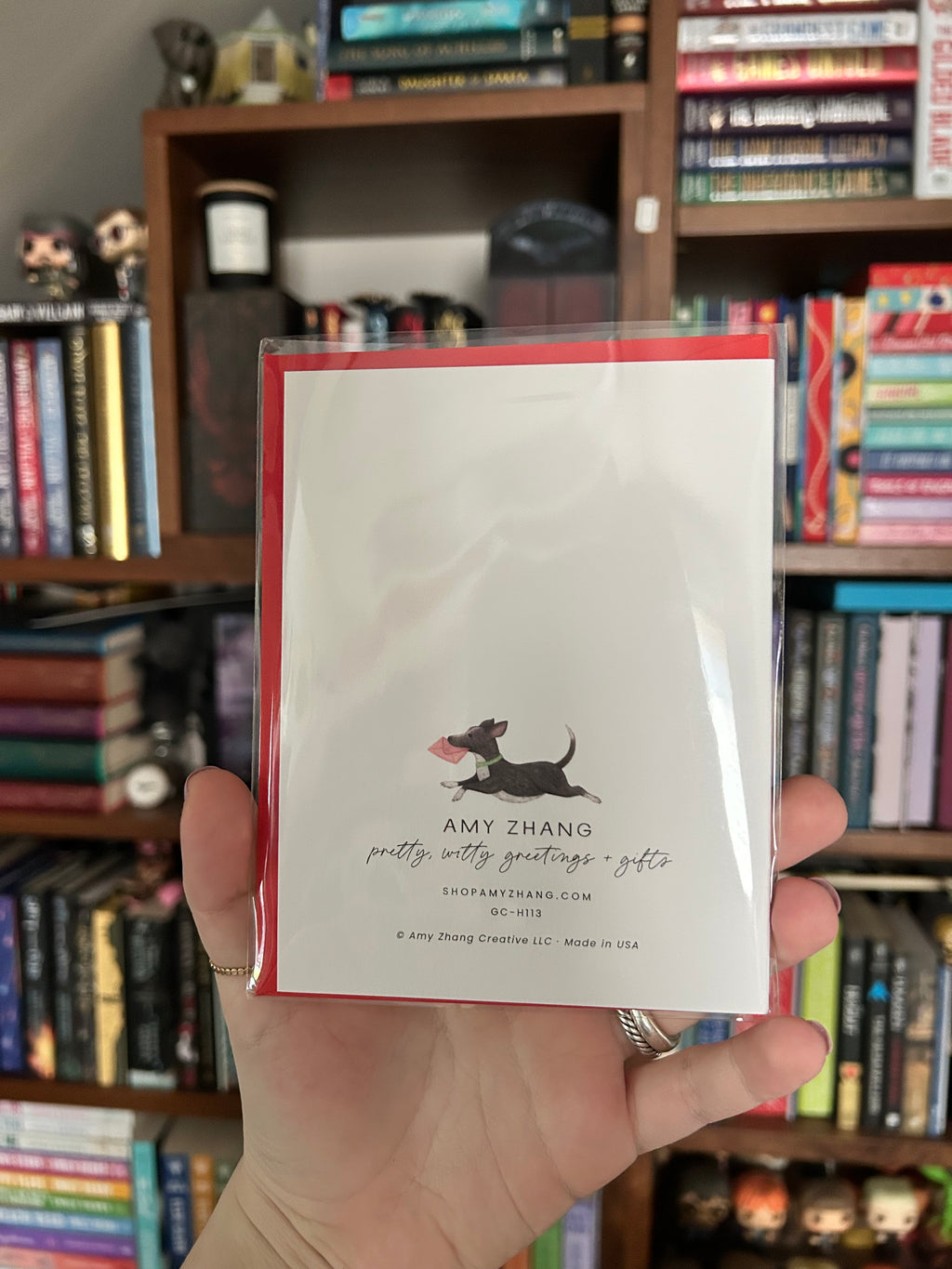 A hand holding a white greeting card with a red envelope and a black dachshund illustration, with a blurred background of bookshelves. 