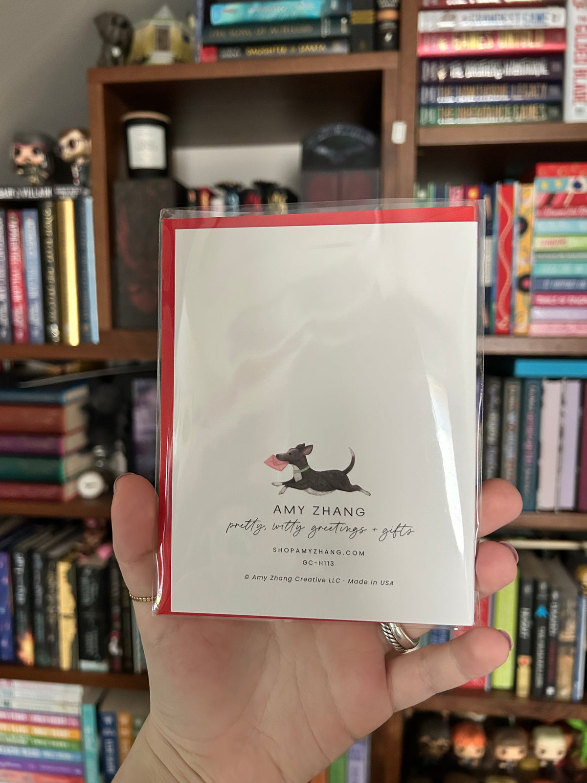 A hand holding a white greeting card with a red envelope and a black dachshund illustration, with a blurred background of bookshelves. 